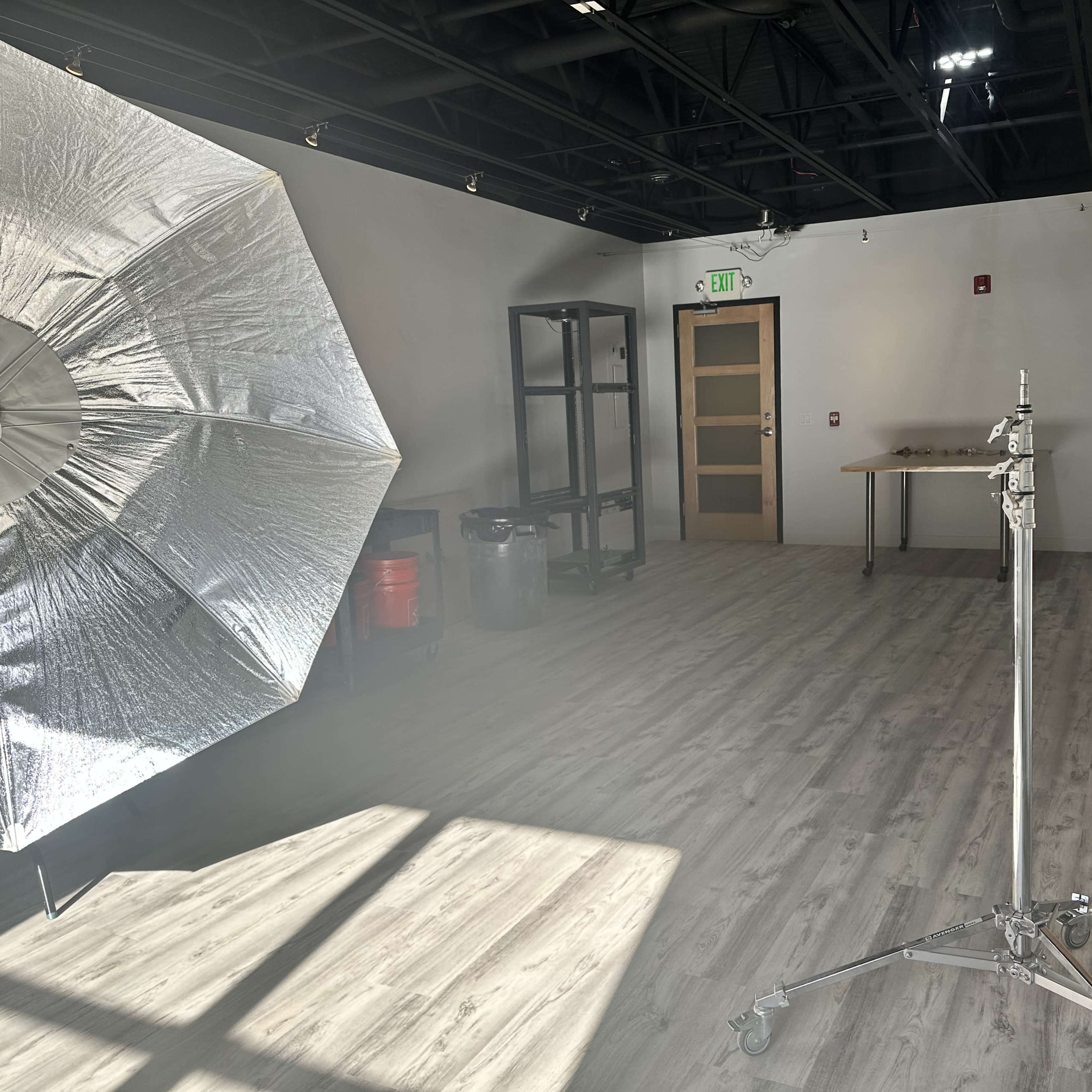 A well-lit studio space with a large reflective umbrella on the left, a light stand, a table, and storage shelves against the wall.