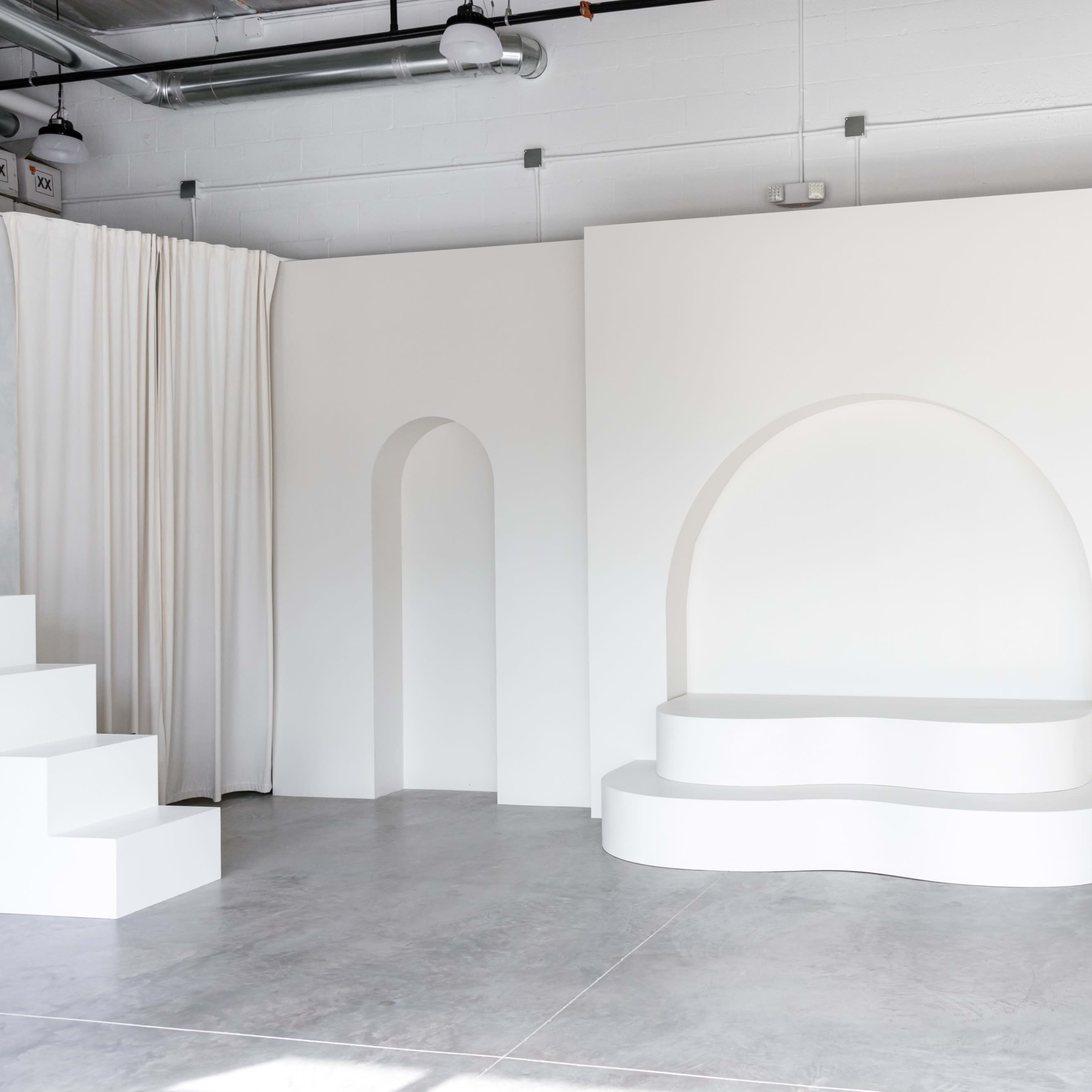 A minimalist, white architectural interior featuring curved and angular walls, an arched niche, and a stepped platform.