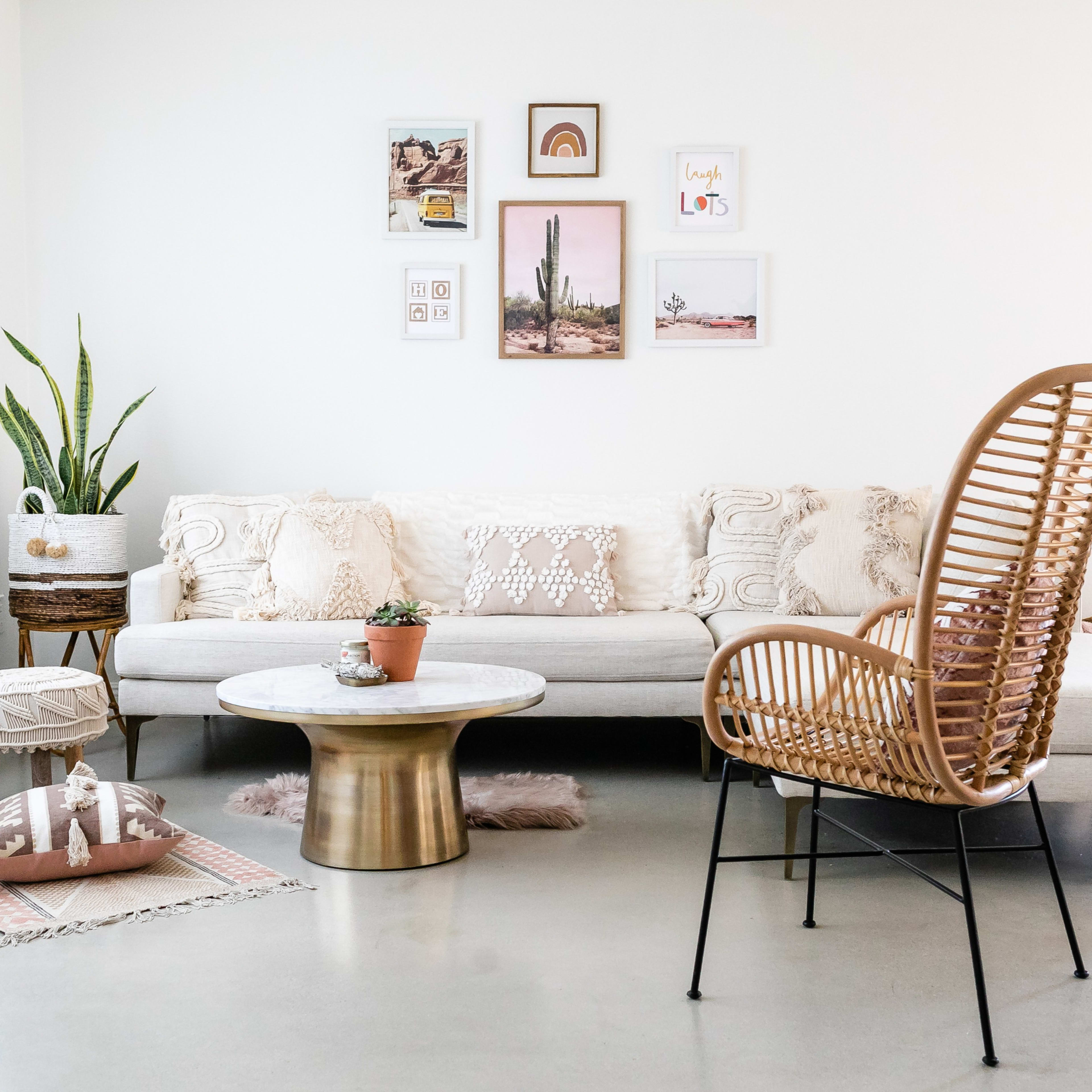 A modern living room features a sectional sofa with decorative pillows, a round coffee table, a rattan chair, and various plants and artwork on the walls.
