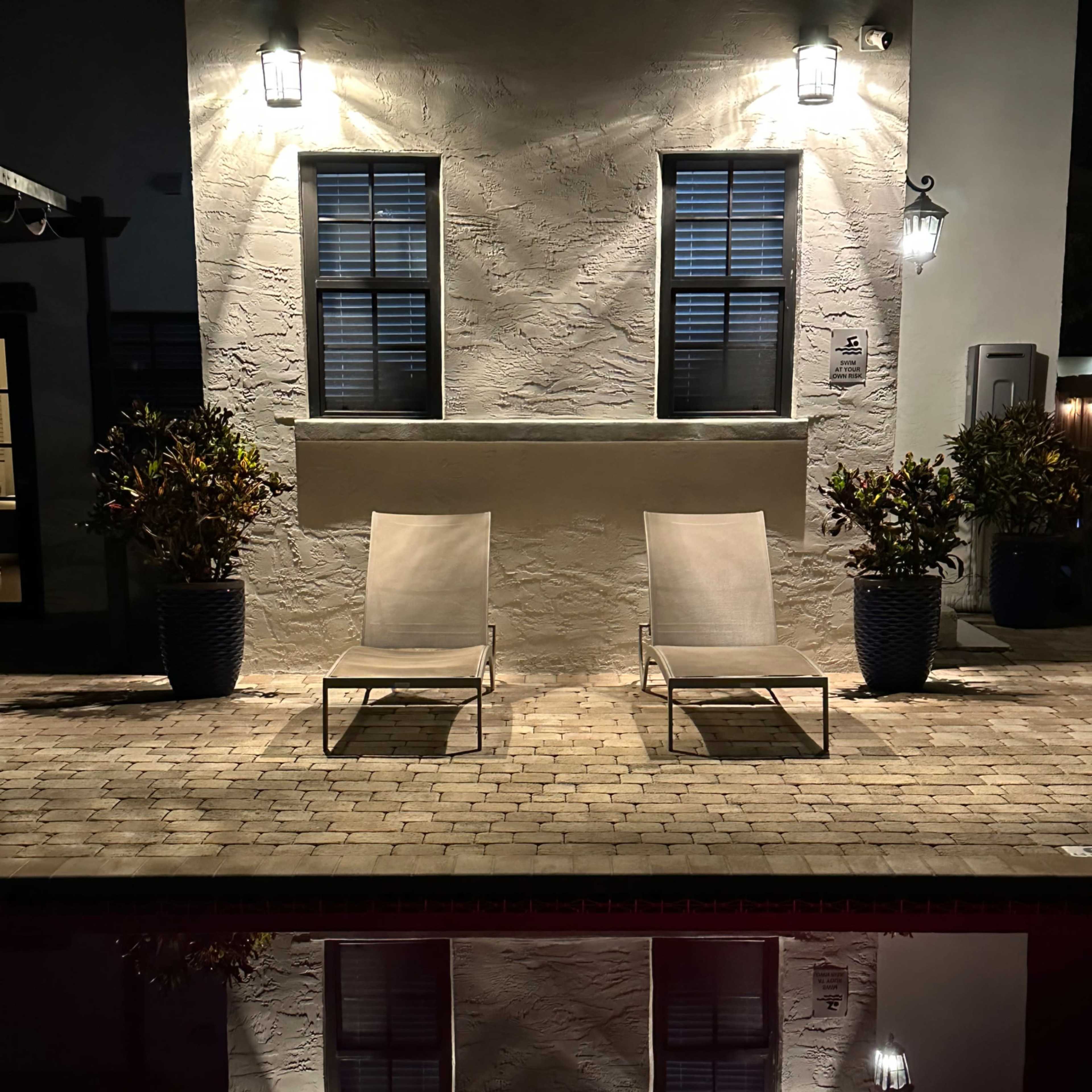 Two lounge chairs sit facing a pool, reflecting the lights and windows of a building at night.