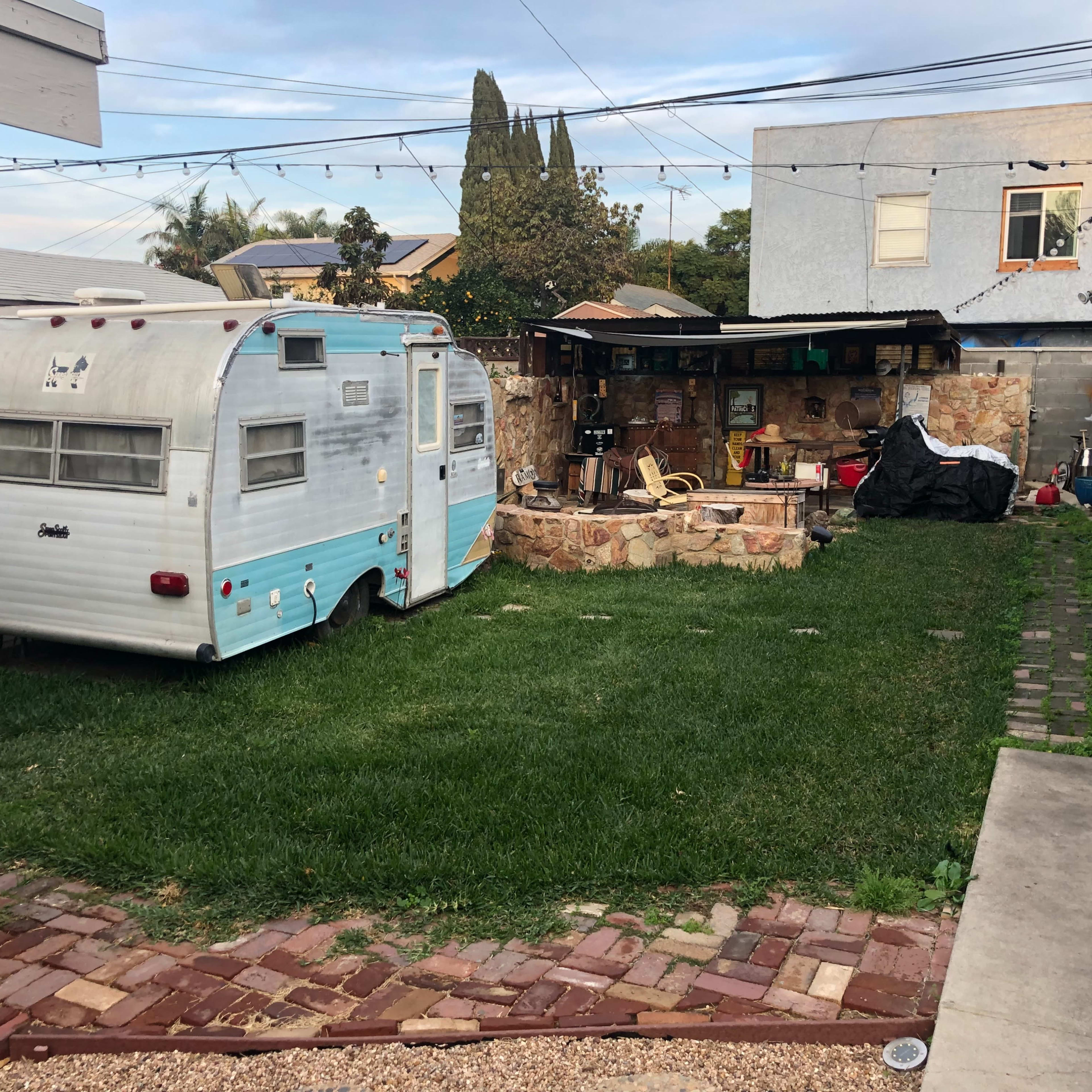 A vintage blue-and-white trailer sits on a grassy yard next to a stone area with outdoor furniture and a covered workspace.