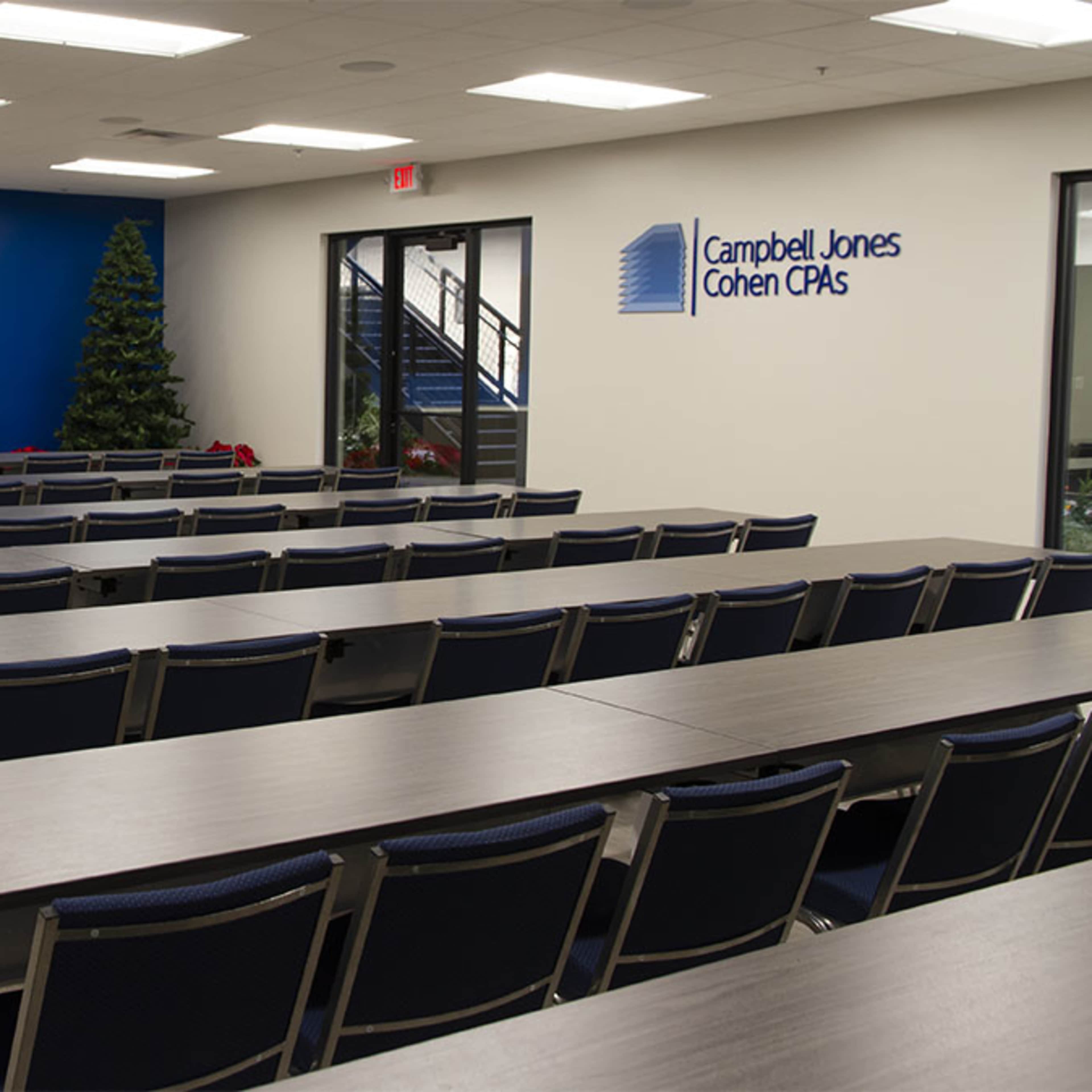 A modern, empty conference room with rows of tables and chairs facing two large screens on a blue accent wall.