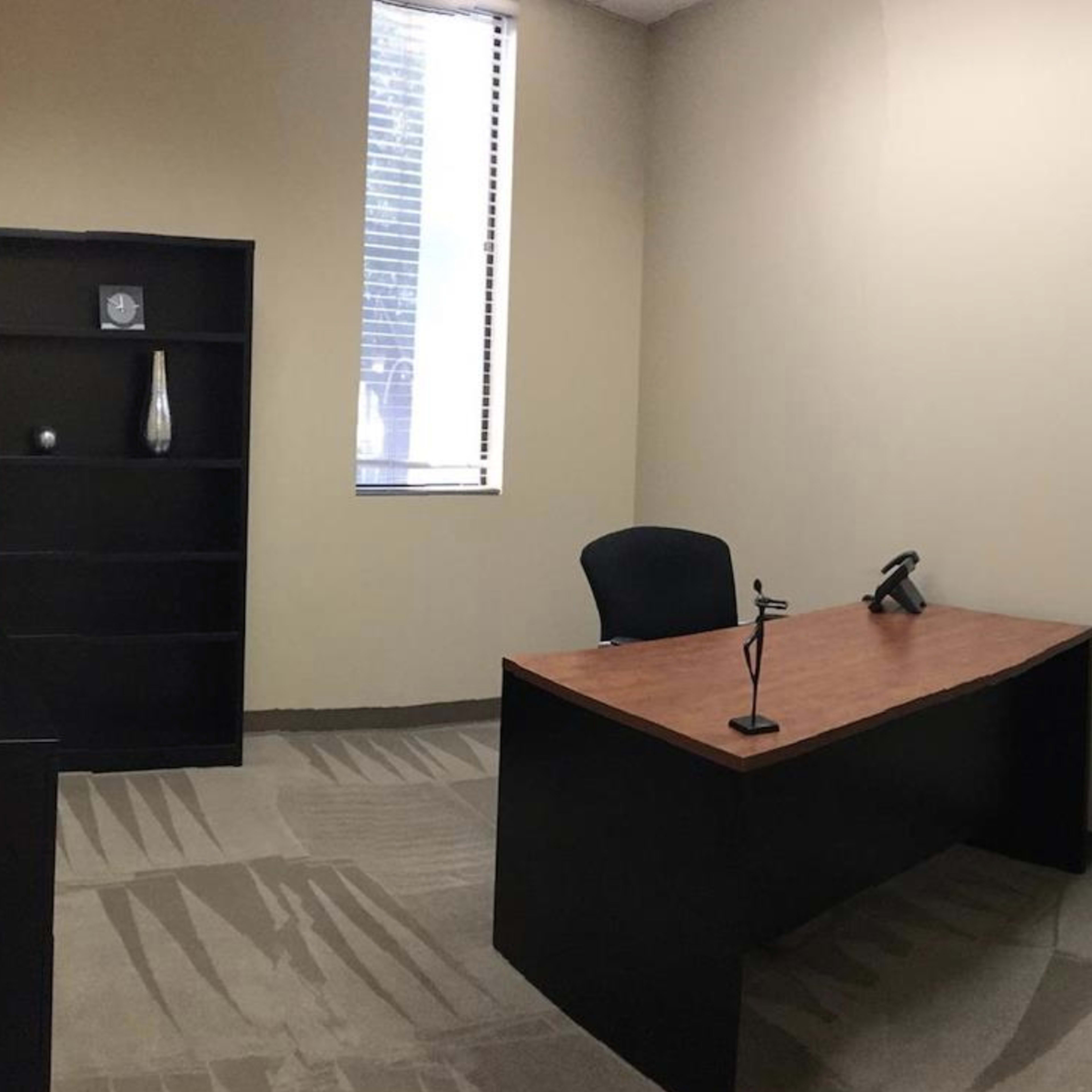 A sparsely furnished office with a black desk and a chair, a shelf against the wall, and a window allowing natural light.
