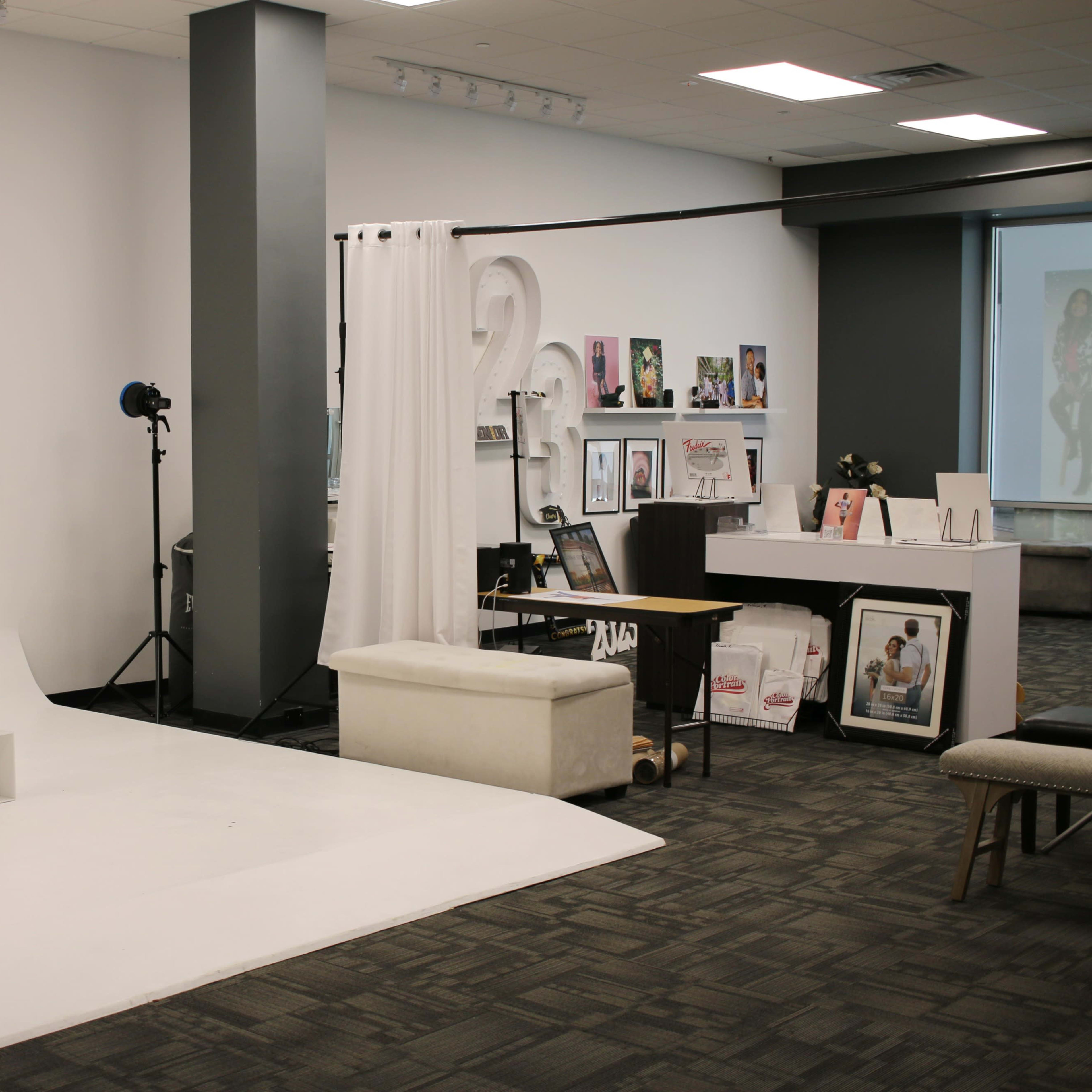 A photography studio features a white backdrop, a large umbrella light, and various displayed photo frames and magazines against a gray wall.