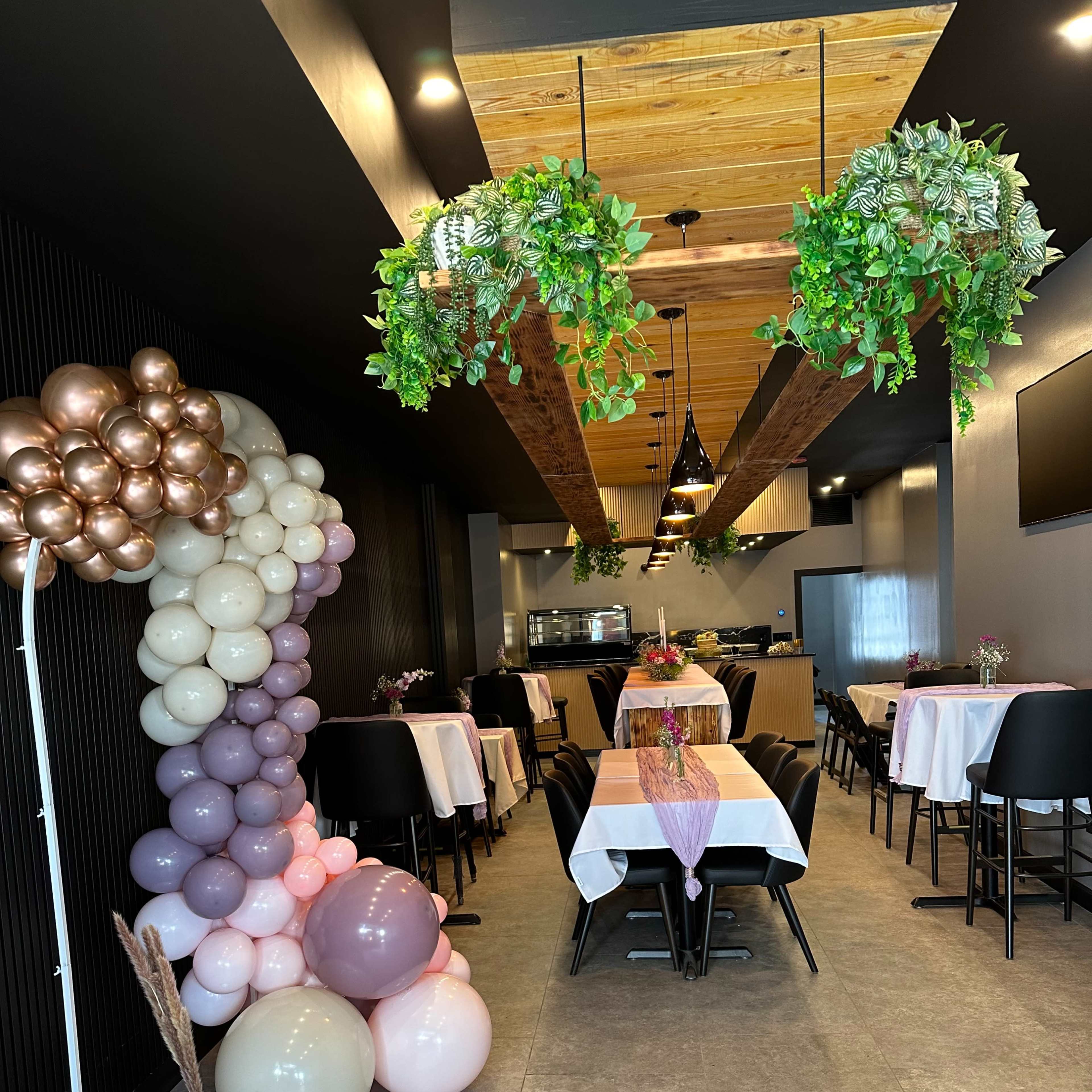 The interior of a decorated restaurant featuring tables with white tablecloths, balloon arrangements in pastel colors, and greenery hanging from the ceiling.