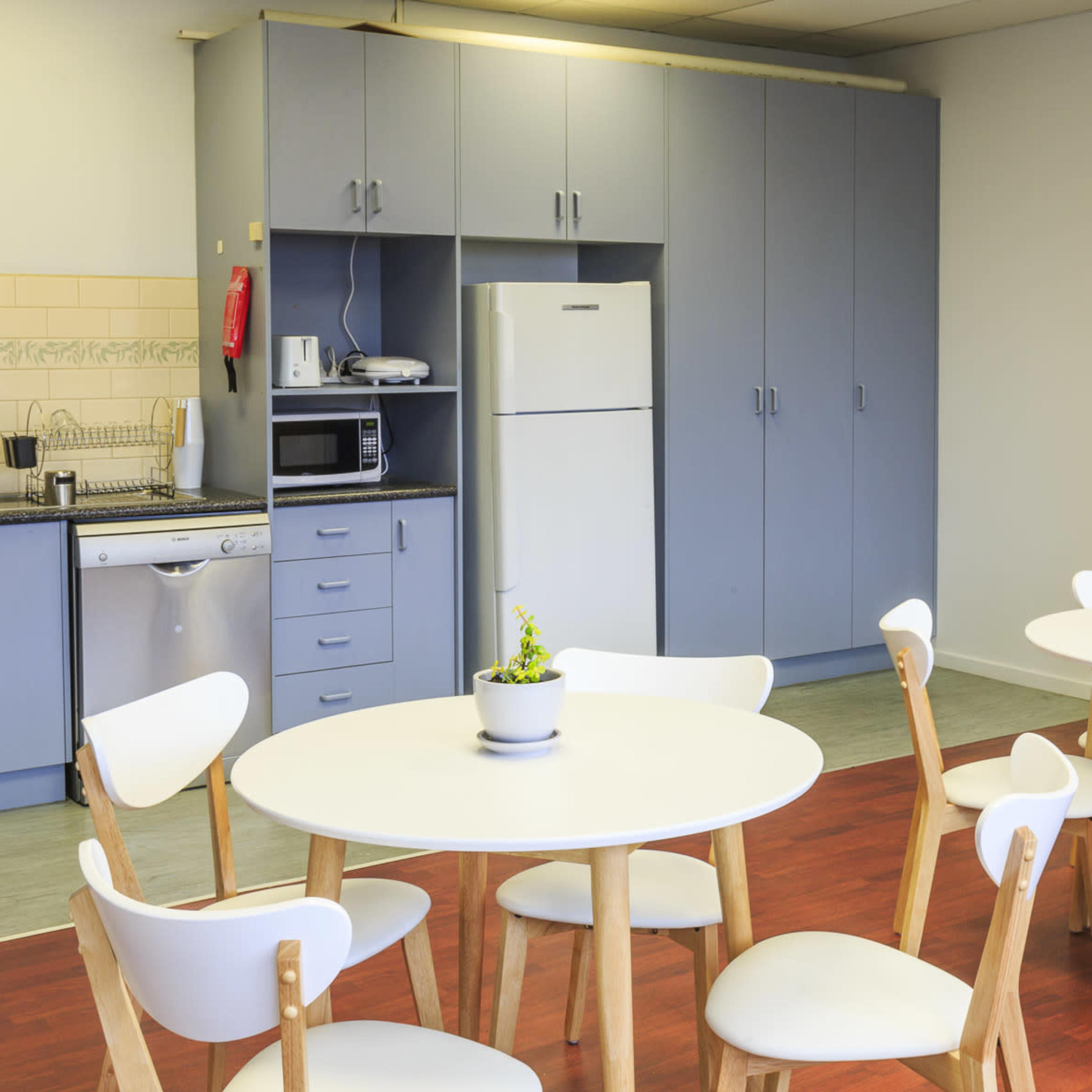 A kitchen area features multiple white round tables surrounded by chairs, with a fridge, microwave, and kitchen cabinets in the background.