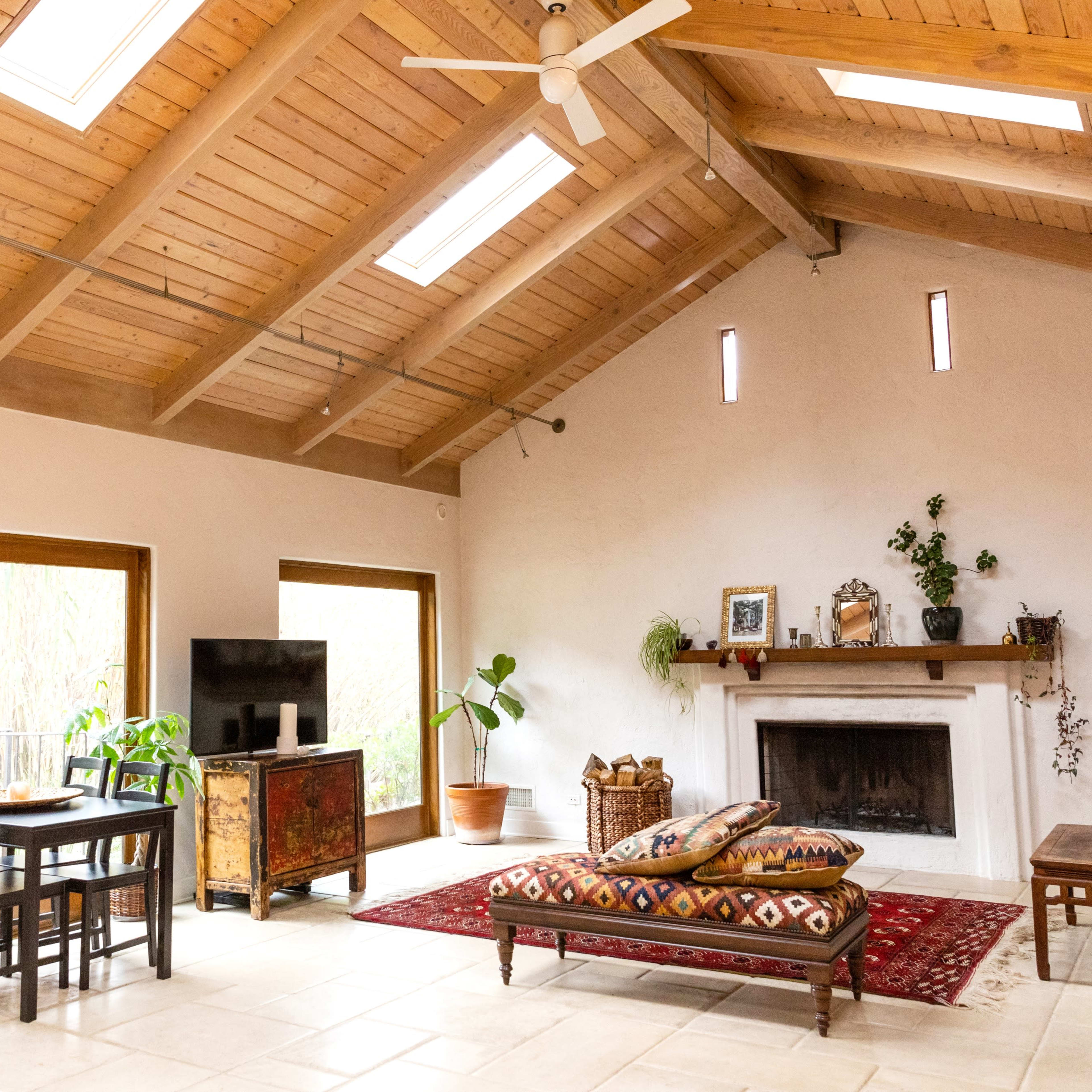 A spacious living area with a high wooden ceiling, a dining table, a television, a sofa, and a fireplace, accented by potted plants and decorative items.