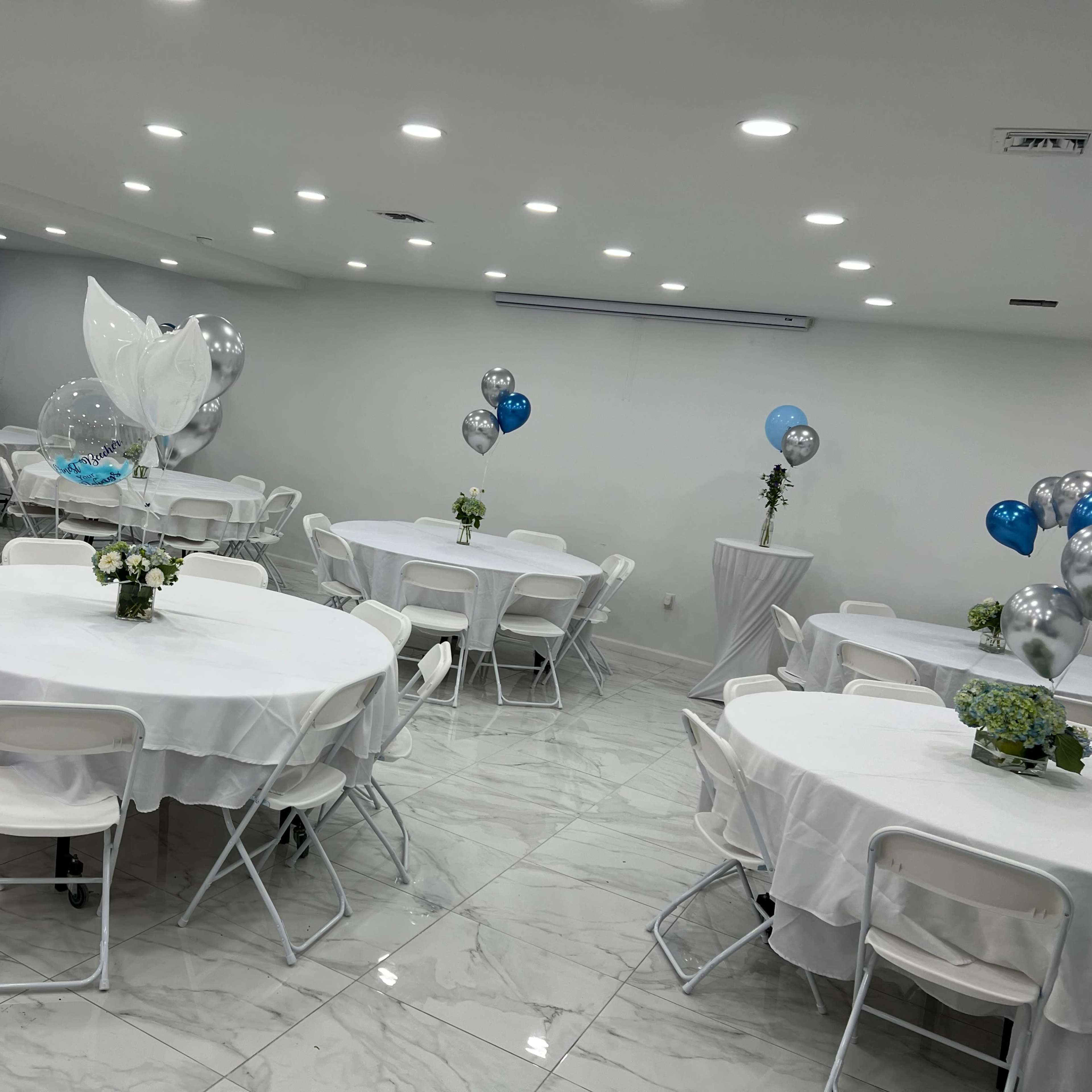 A brightly lit event space with several round tables covered in white tablecloths, each adorned with centerpieces, and decorated with balloons in blue and silver.