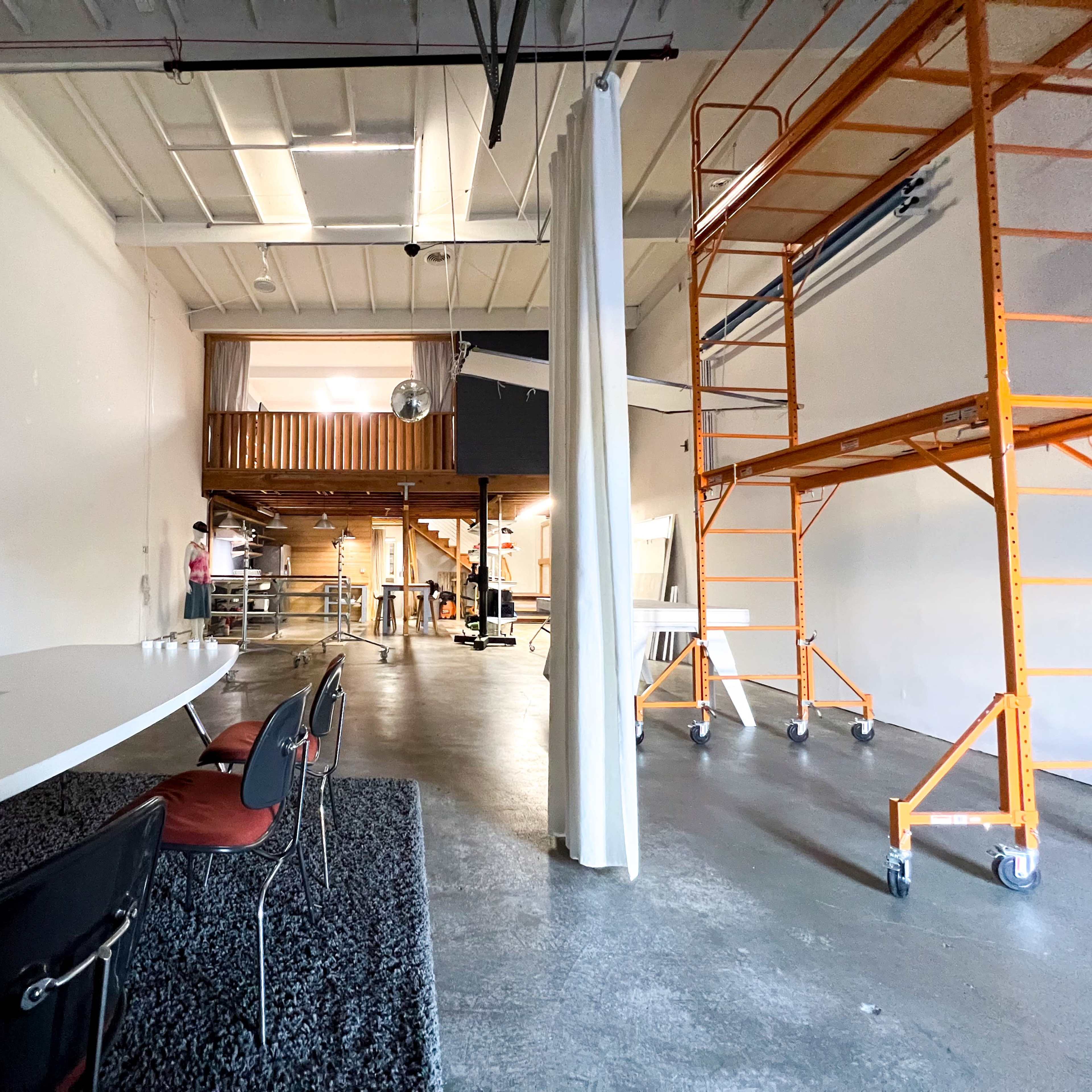 A spacious, industrial-style room featuring a long conference table, a scaffolding structure, and a second-level loft area.