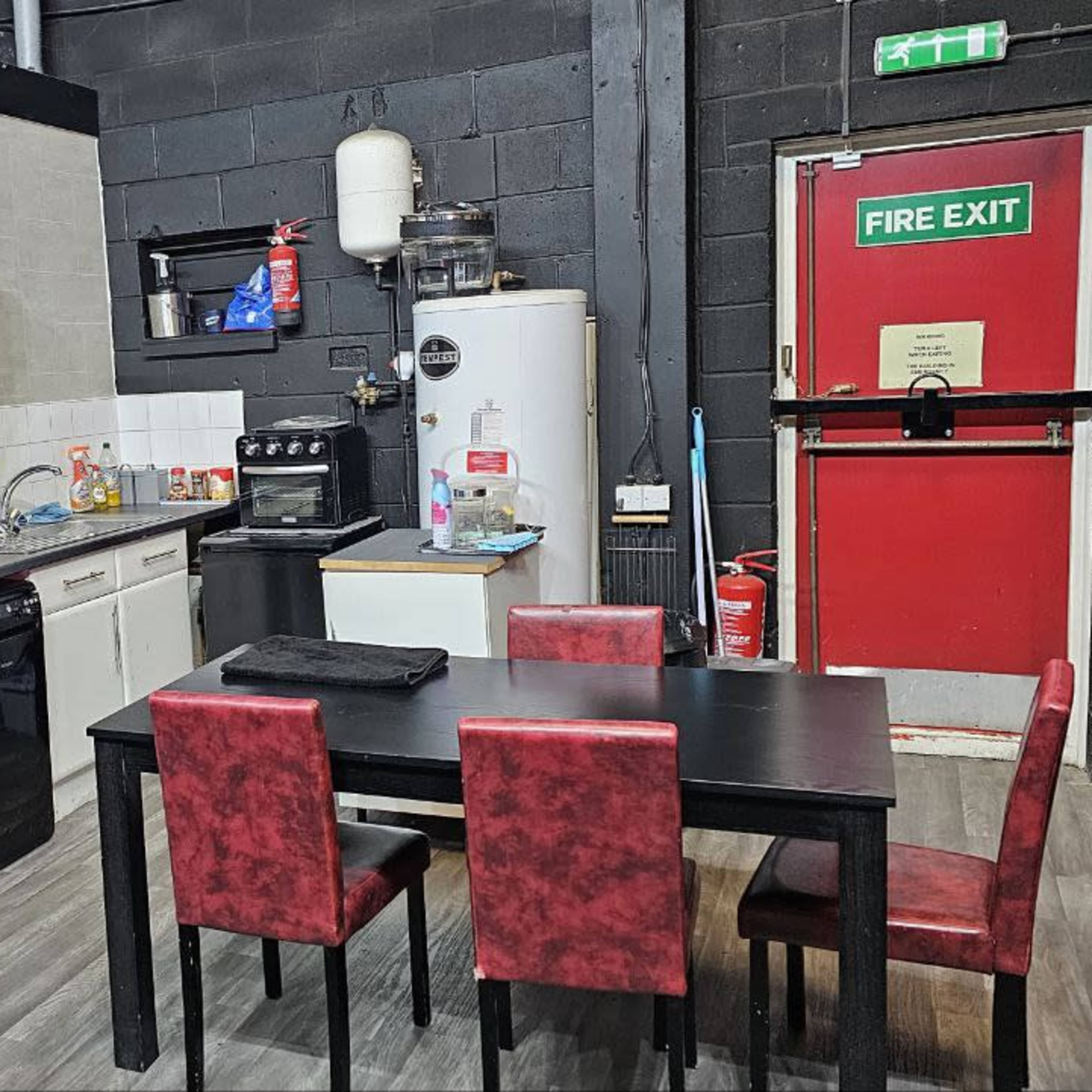 A kitchen area with a black table and red chairs, a refrigerator, a stove, and a fire exit door.