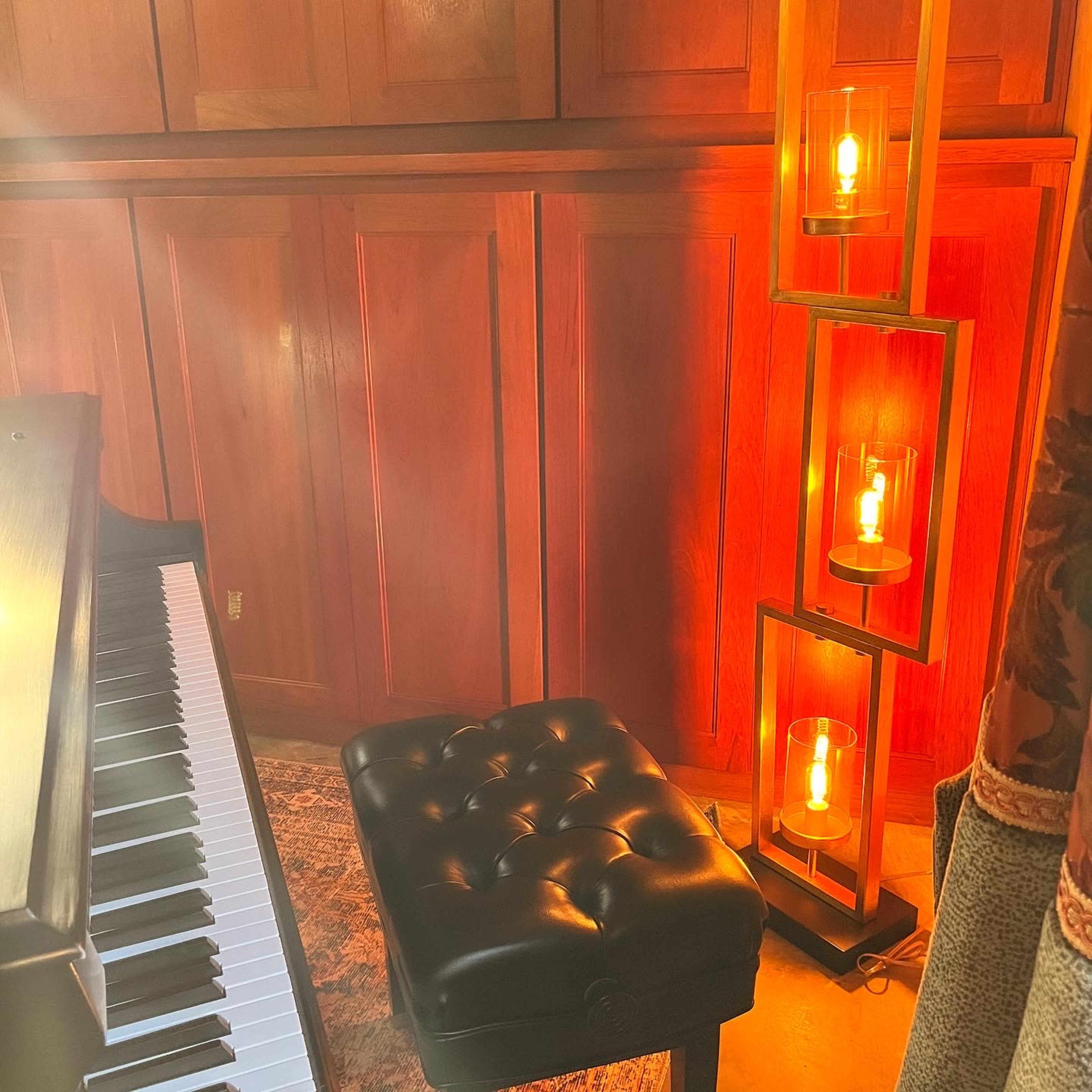 A black piano bench sits beside a grand piano near a tall lamp with glowing light bulbs against a wooden wall.