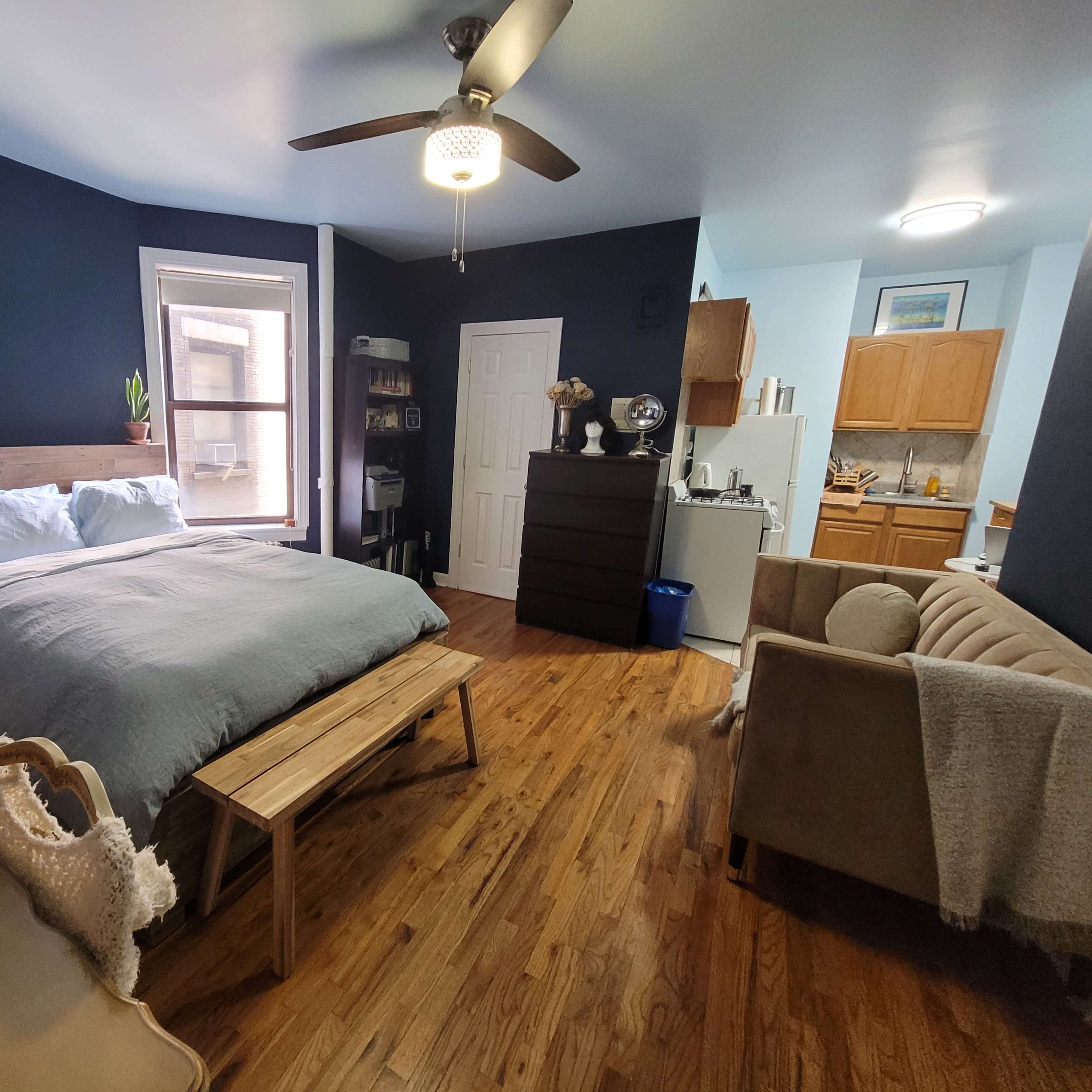 A well-furnished studio apartment featuring a bed, a seating area, a kitchenette, and hardwood floors.