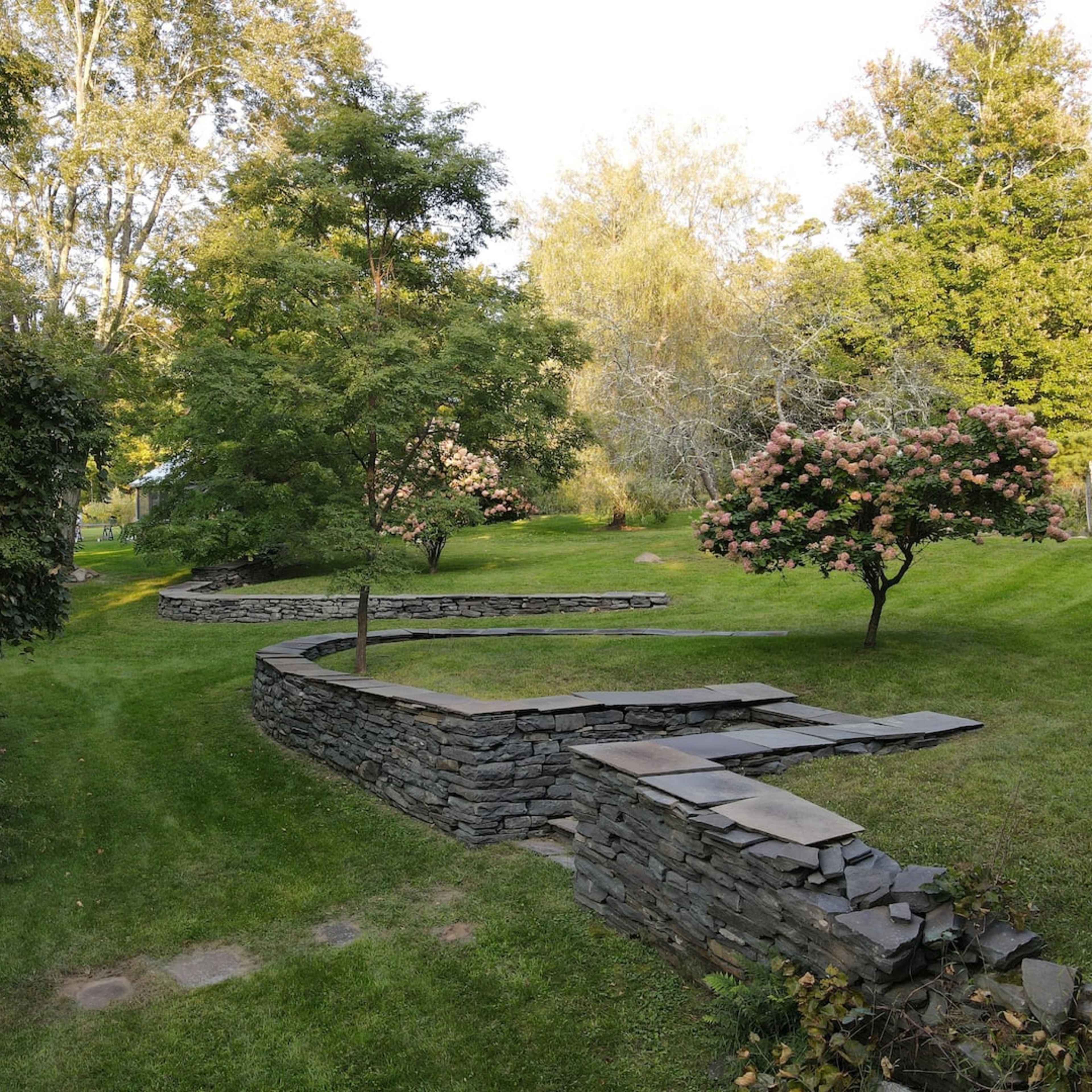 A landscaped garden featuring curved stone walls, a pathway, and trees with blooming flowers.
