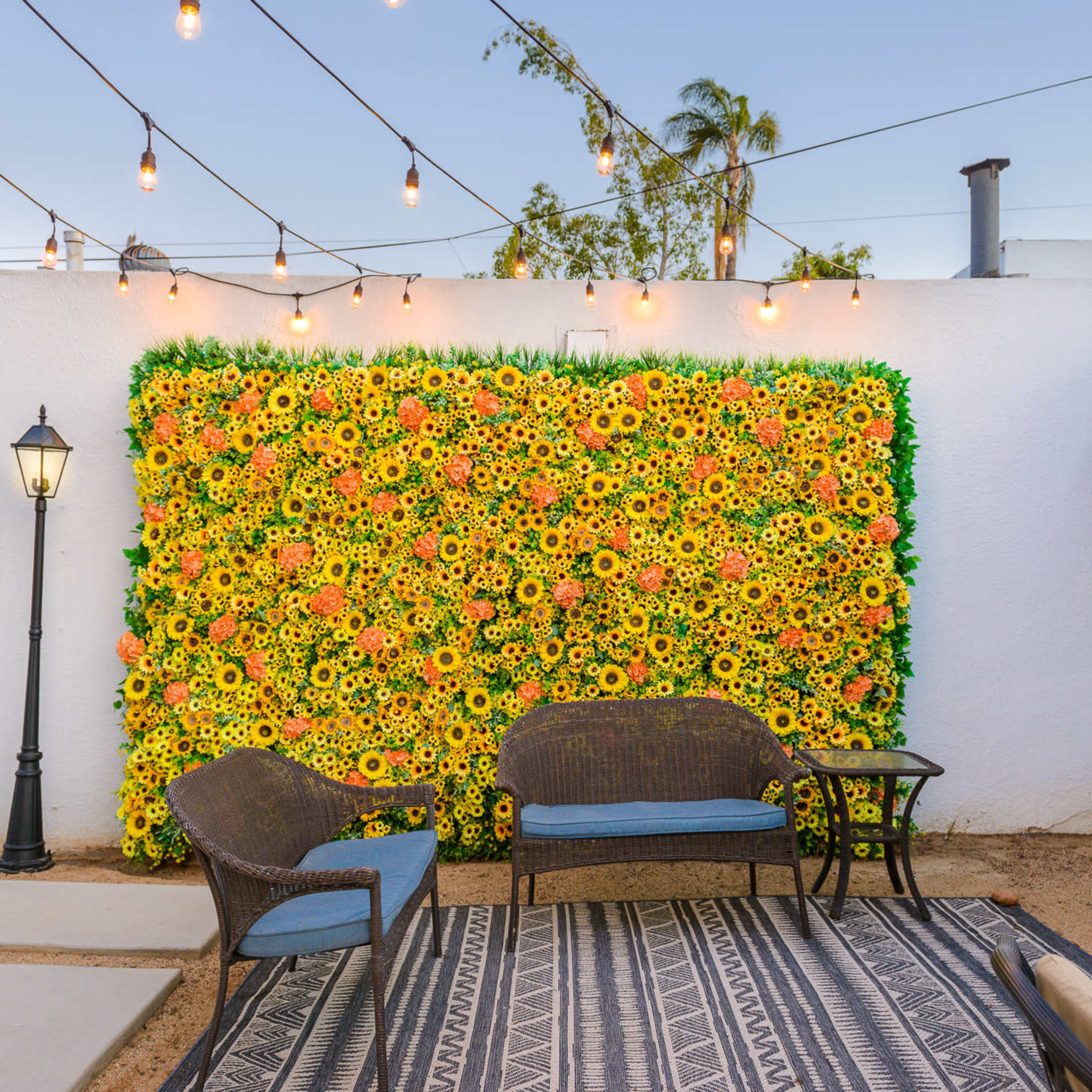 A decorated outdoor space features a vibrant floral wall of yellow and orange flowers, paired with rattan seating and string lights overhead.