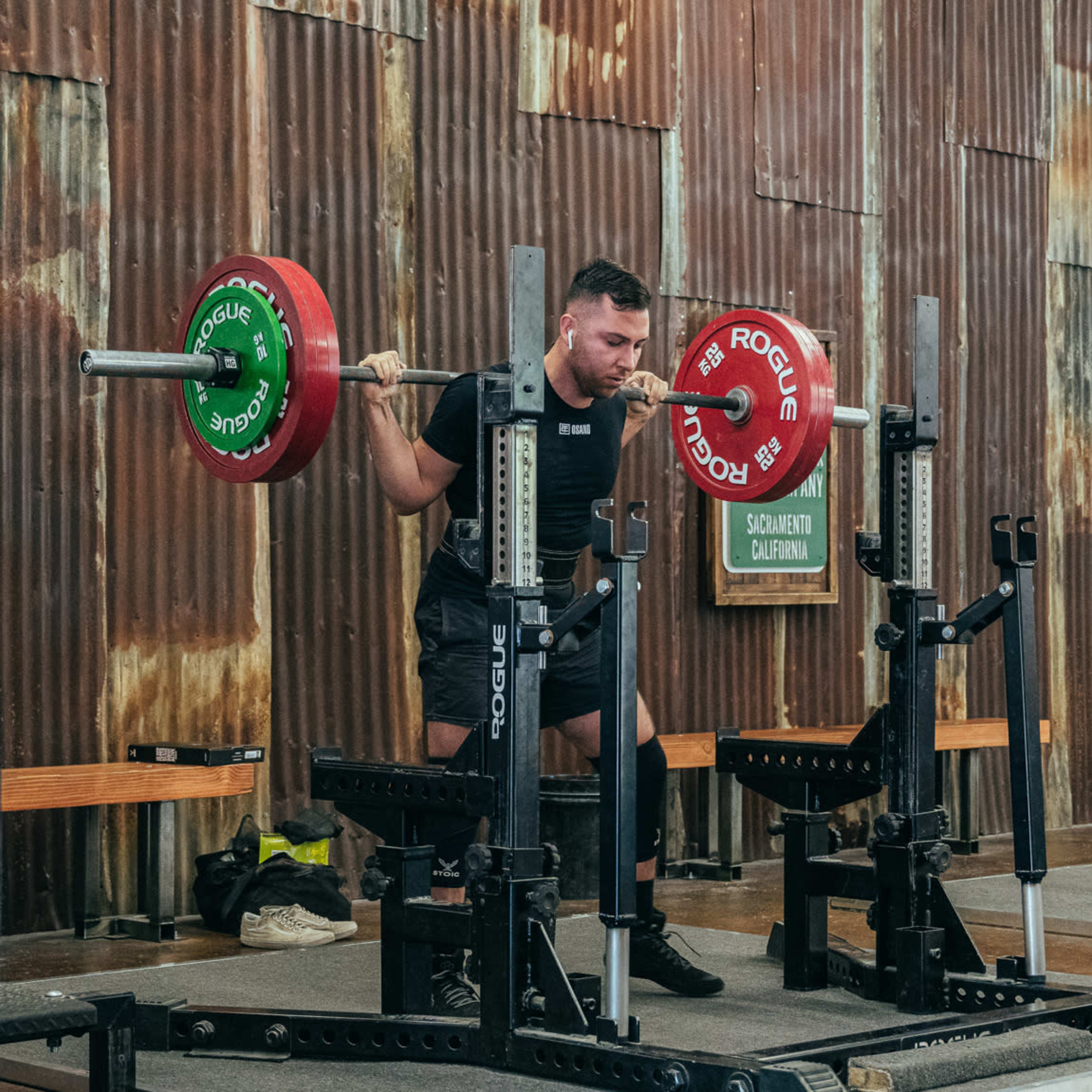 A person is performing a squat with a barbell loaded with weights in a gym setting with metal walls and equipment.