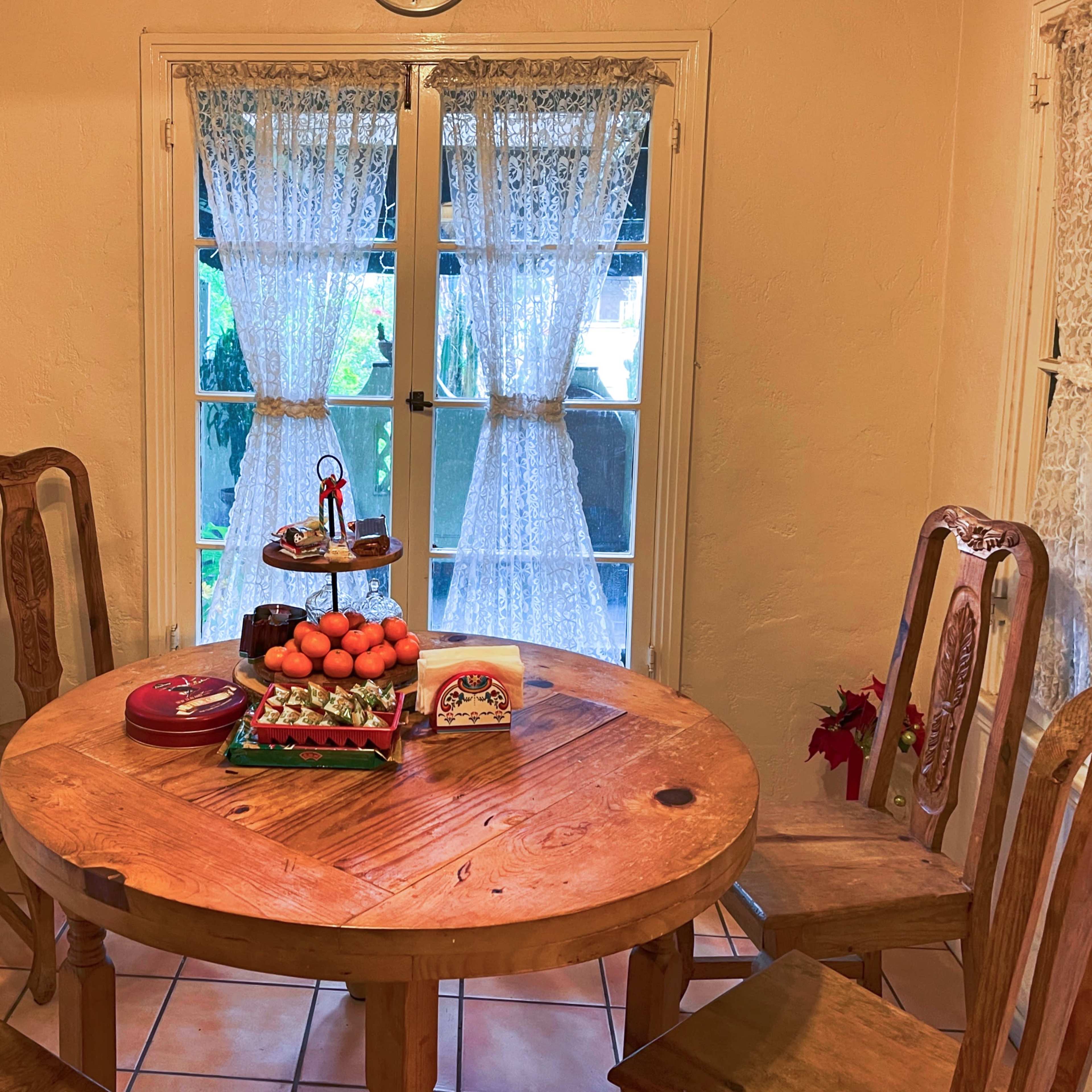 A wooden table set with fruit and decorative items is surrounded by four chairs in a bright room with lace curtains.