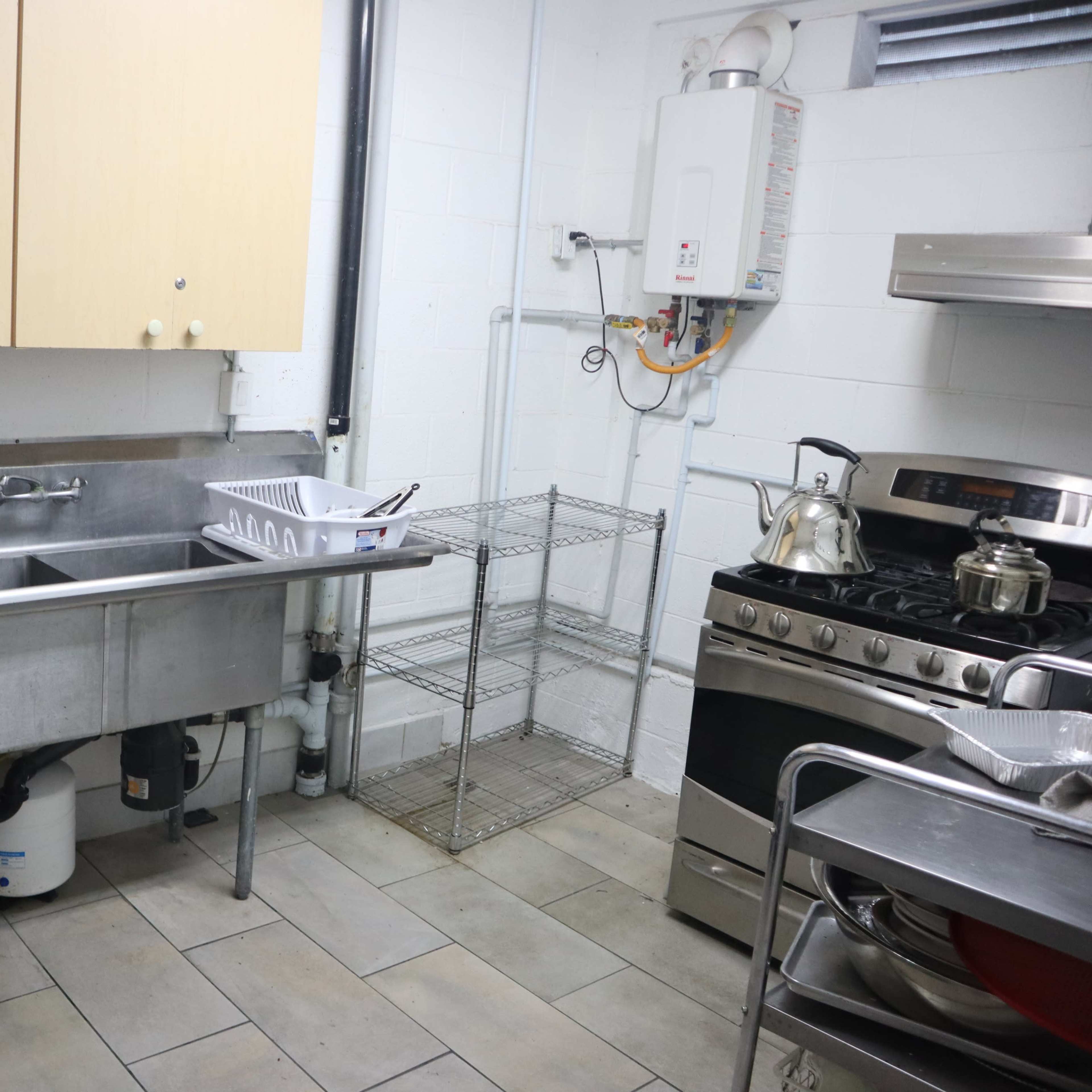 A commercial kitchen features a stainless steel sink, a gas stove, a metal shelf, and wooden cabinets against the wall.