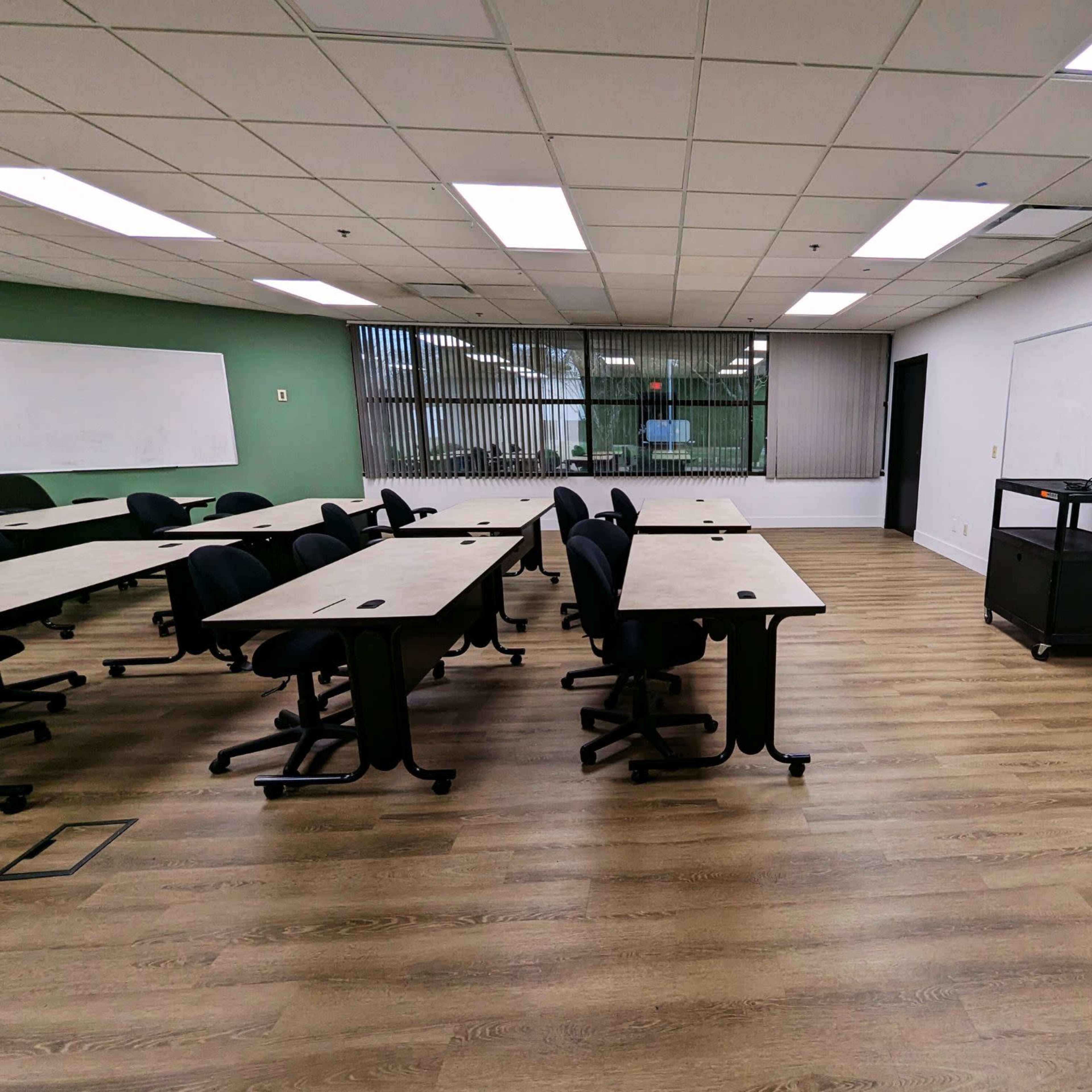 A classroom features several tables arranged with chairs, large windows overlooking greenery, and whiteboards on two walls.