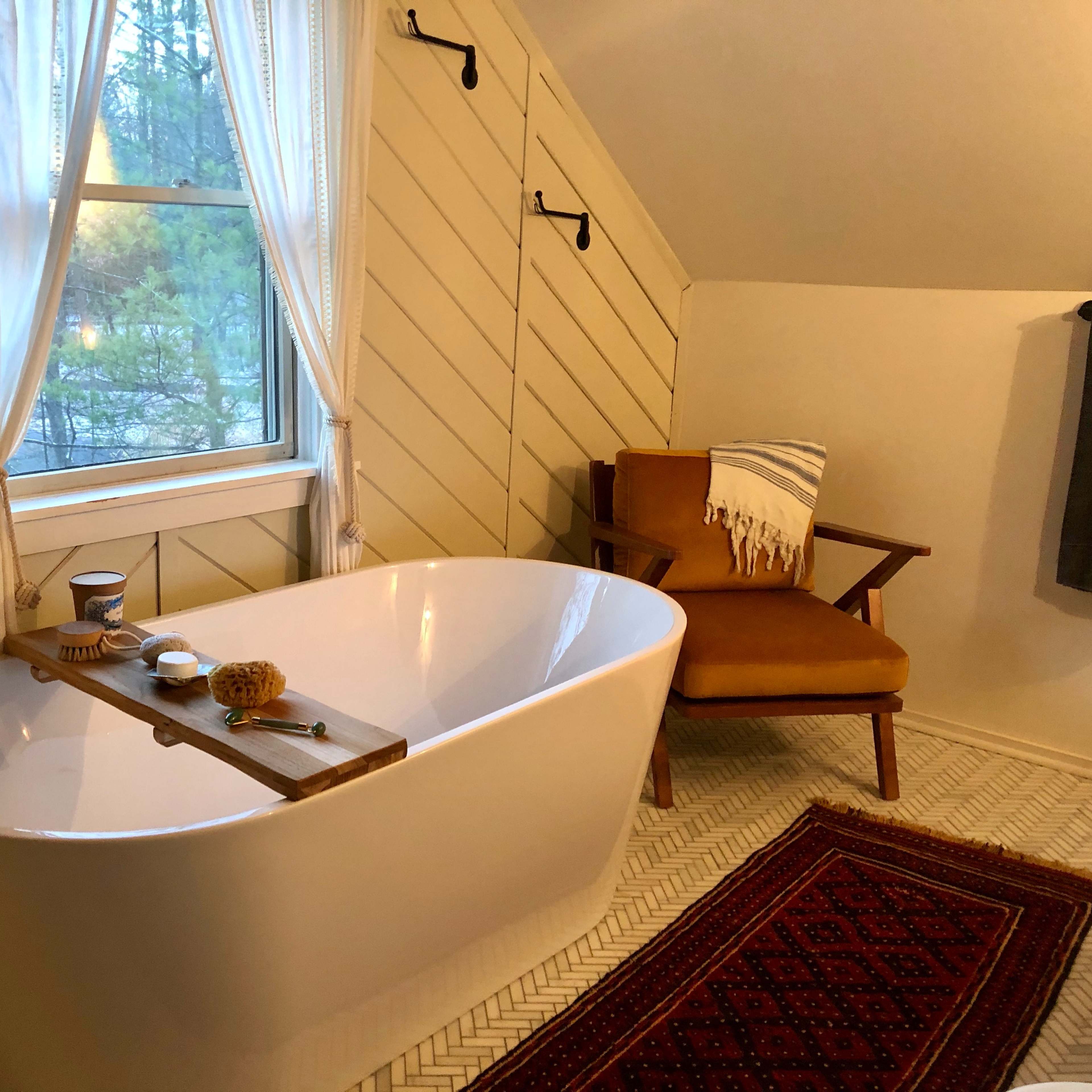 A modern bathroom with a freestanding bathtub, a wooden chair, and natural light coming through a window.