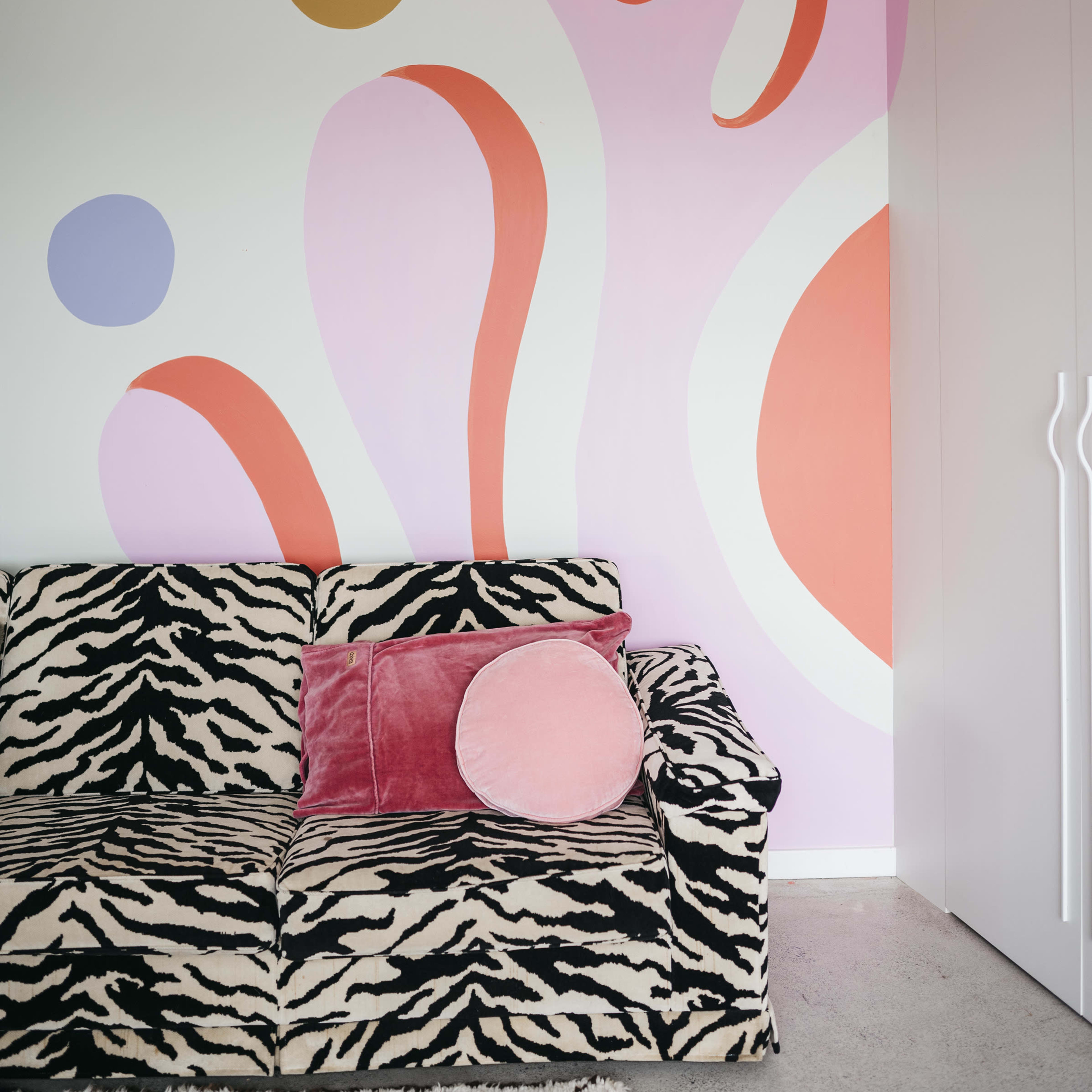 A zebra-striped sofa with a pink pillow against a wall featuring abstract pastel-colored shapes and patterns.