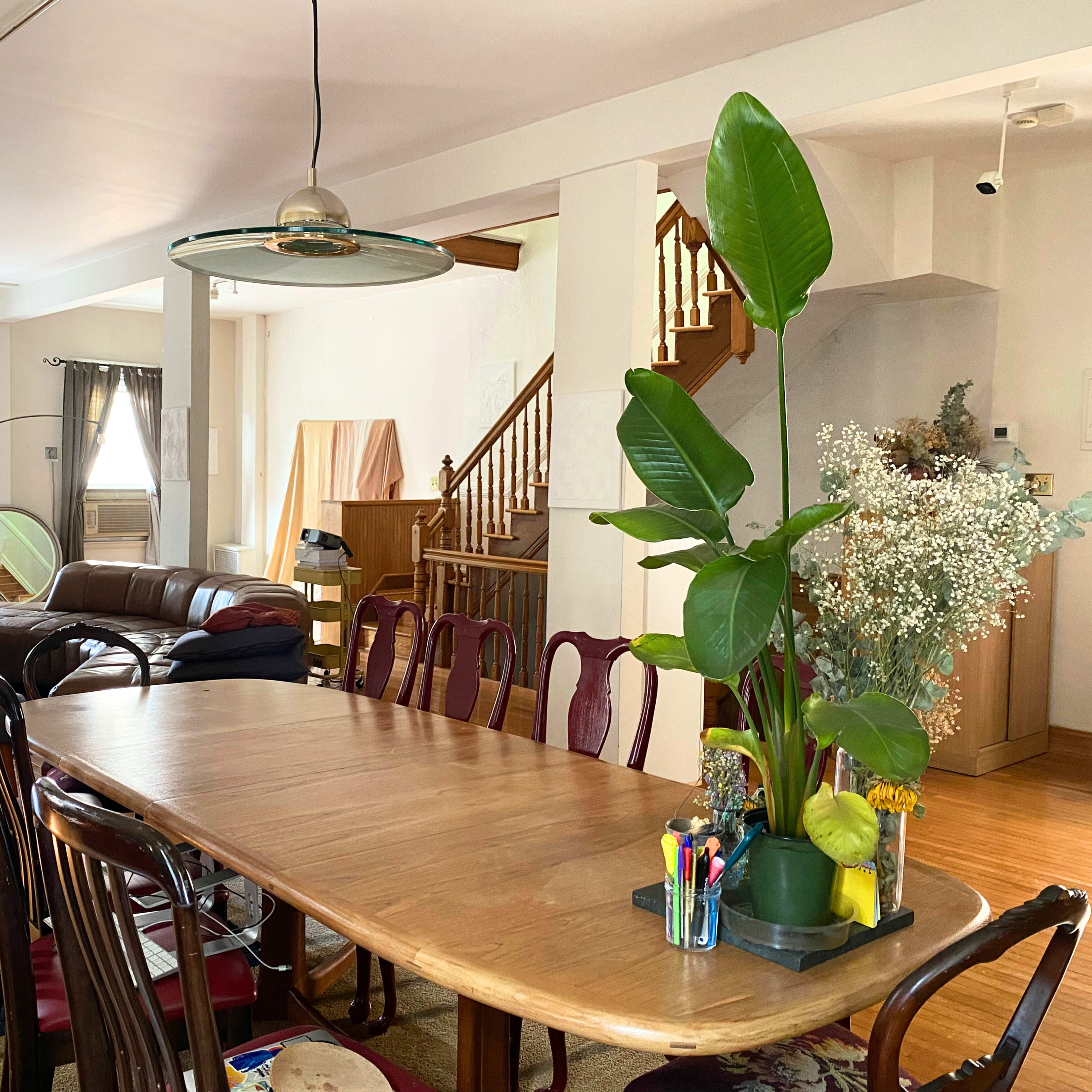A spacious dining area with a long wooden table, surrounded by chairs, and connected to a living space featuring a sofa and a staircase in the background.