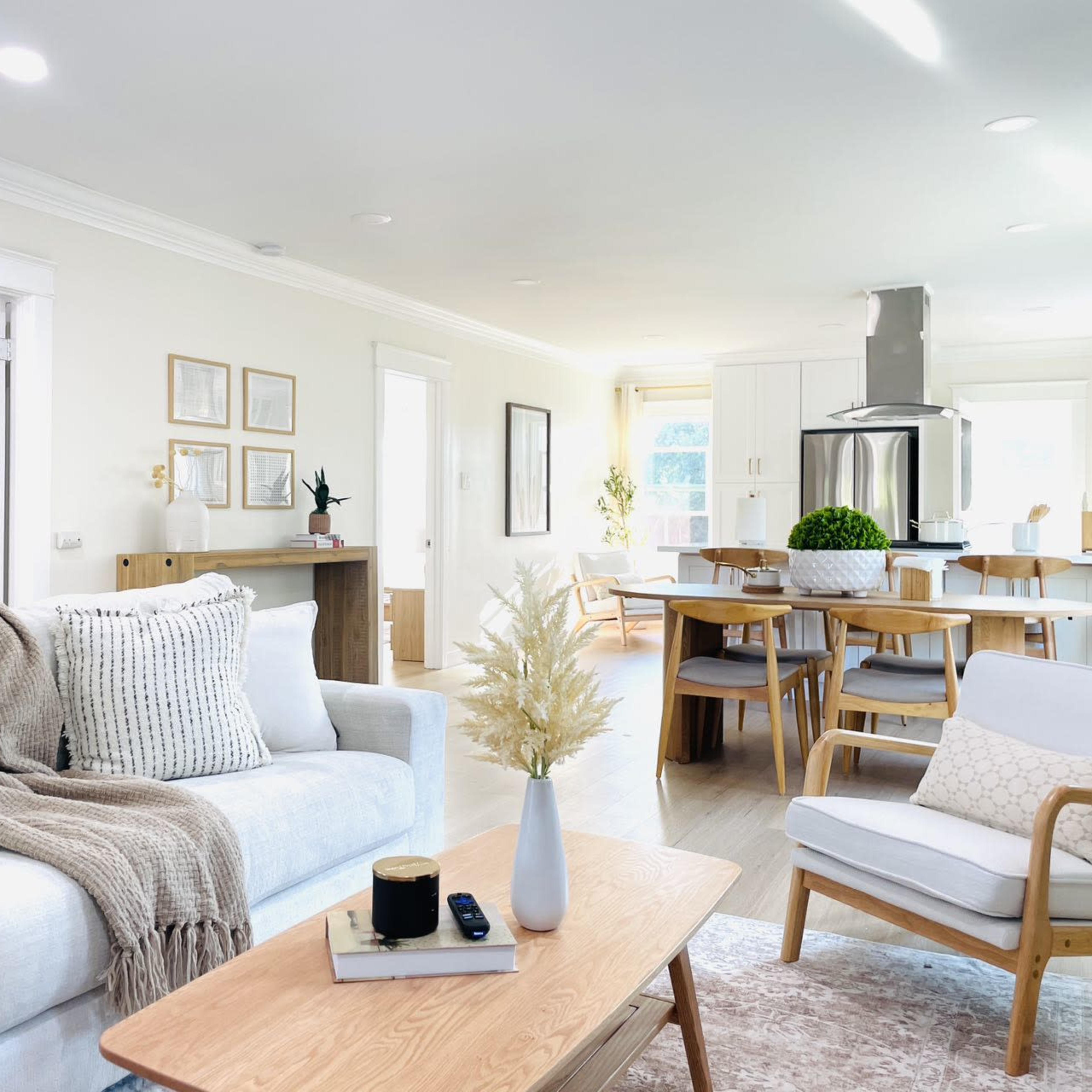 A modern living room features a light-colored sofa with decorative pillows, a wooden coffee table, and an open layout leading to a dining area with a large table and kitchen in the background.