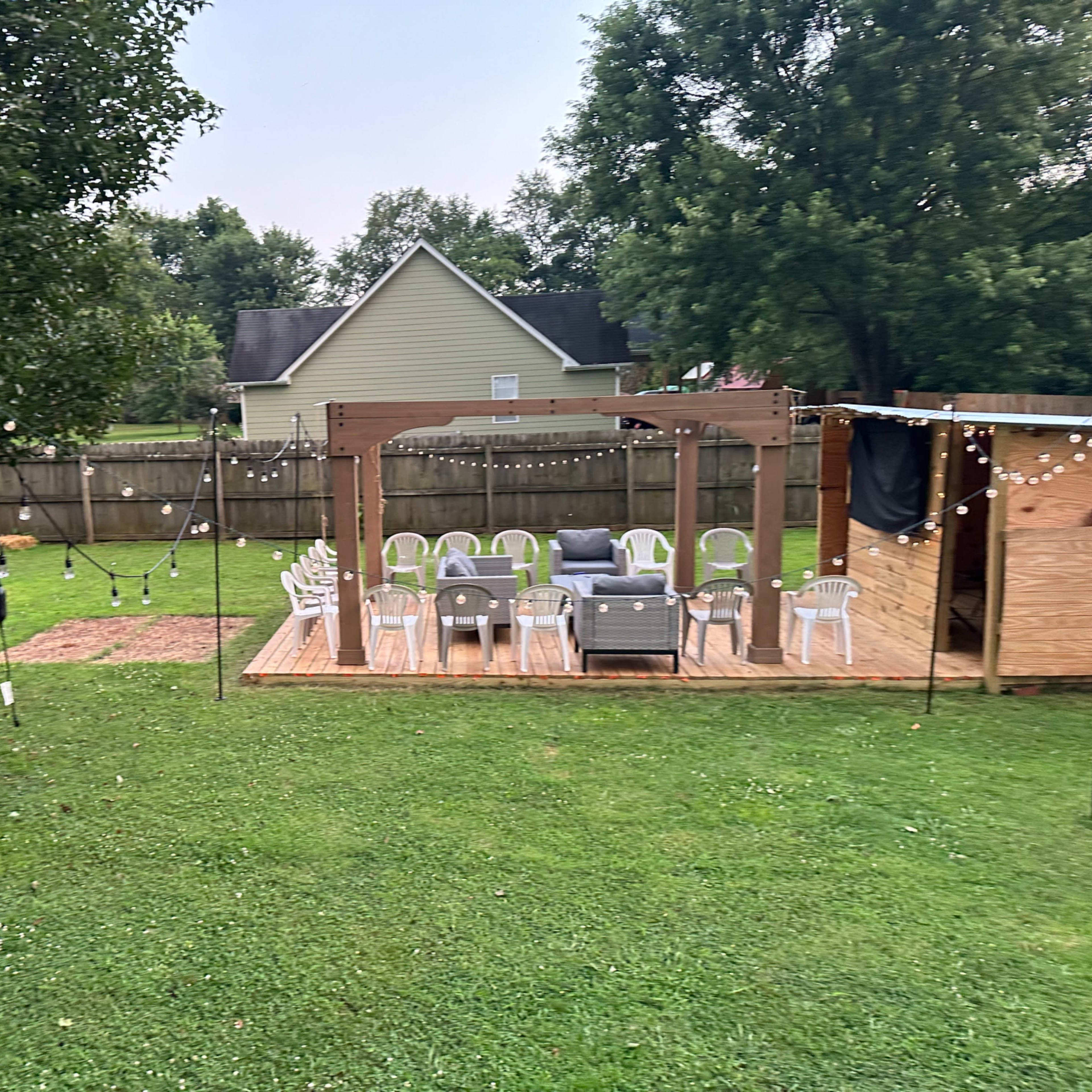 A grassy backyard with a wooden deck containing chairs and a couch, surrounded by a fence and trees.