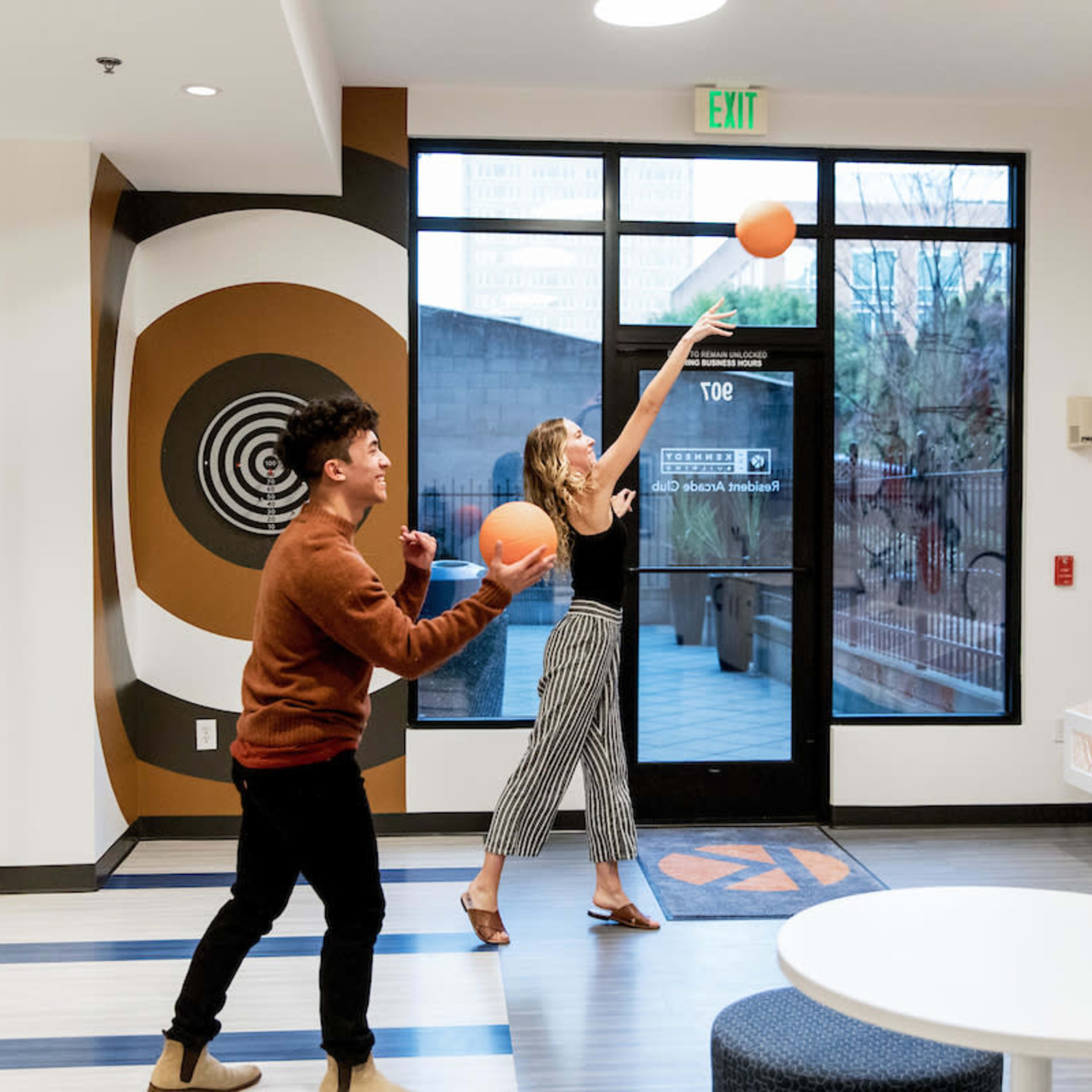 Two people are playing basketball in a recreational room with a large window and decorative wall graphics.