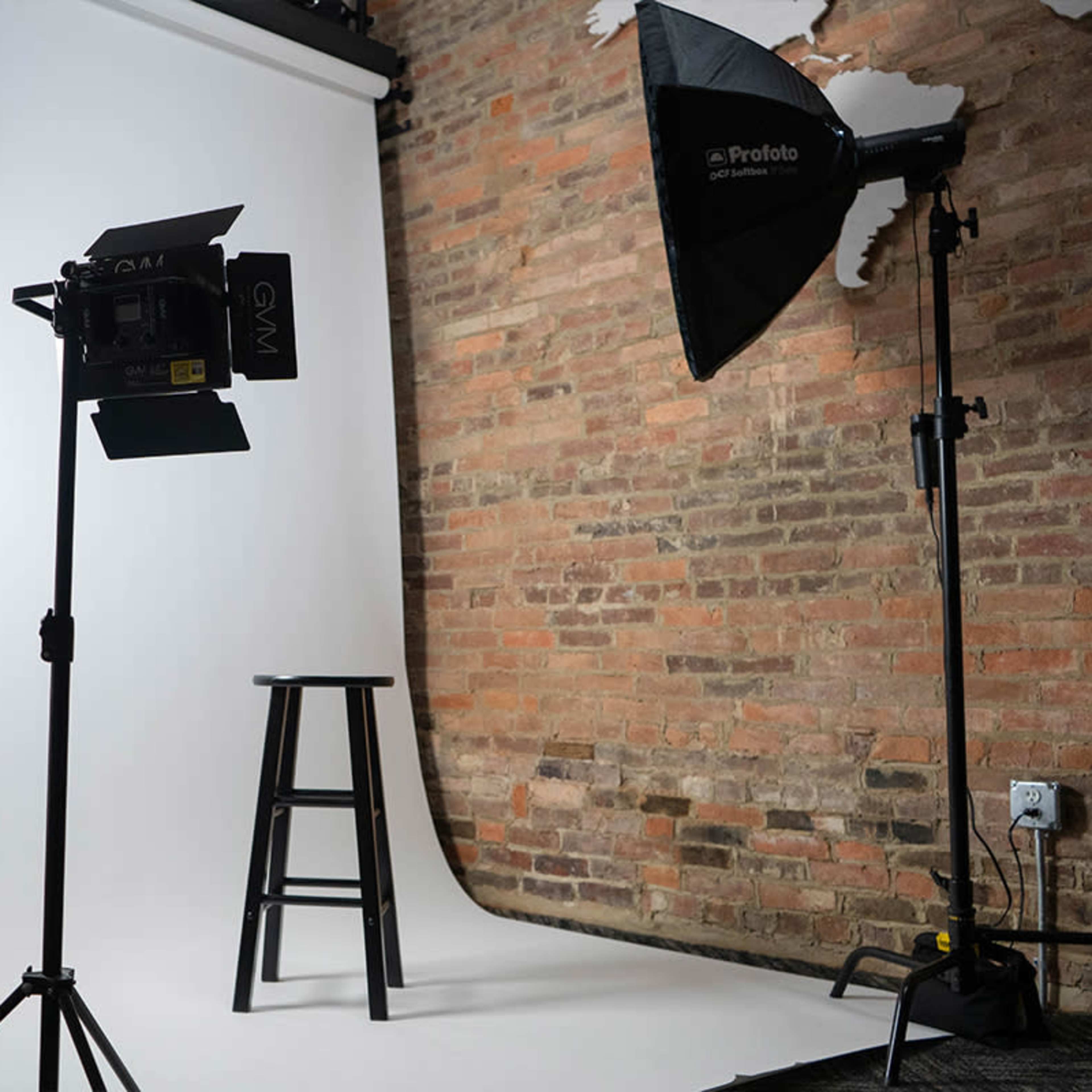 A photography studio setup featuring a white backdrop, two softbox lights, and a black stool positioned in the center.