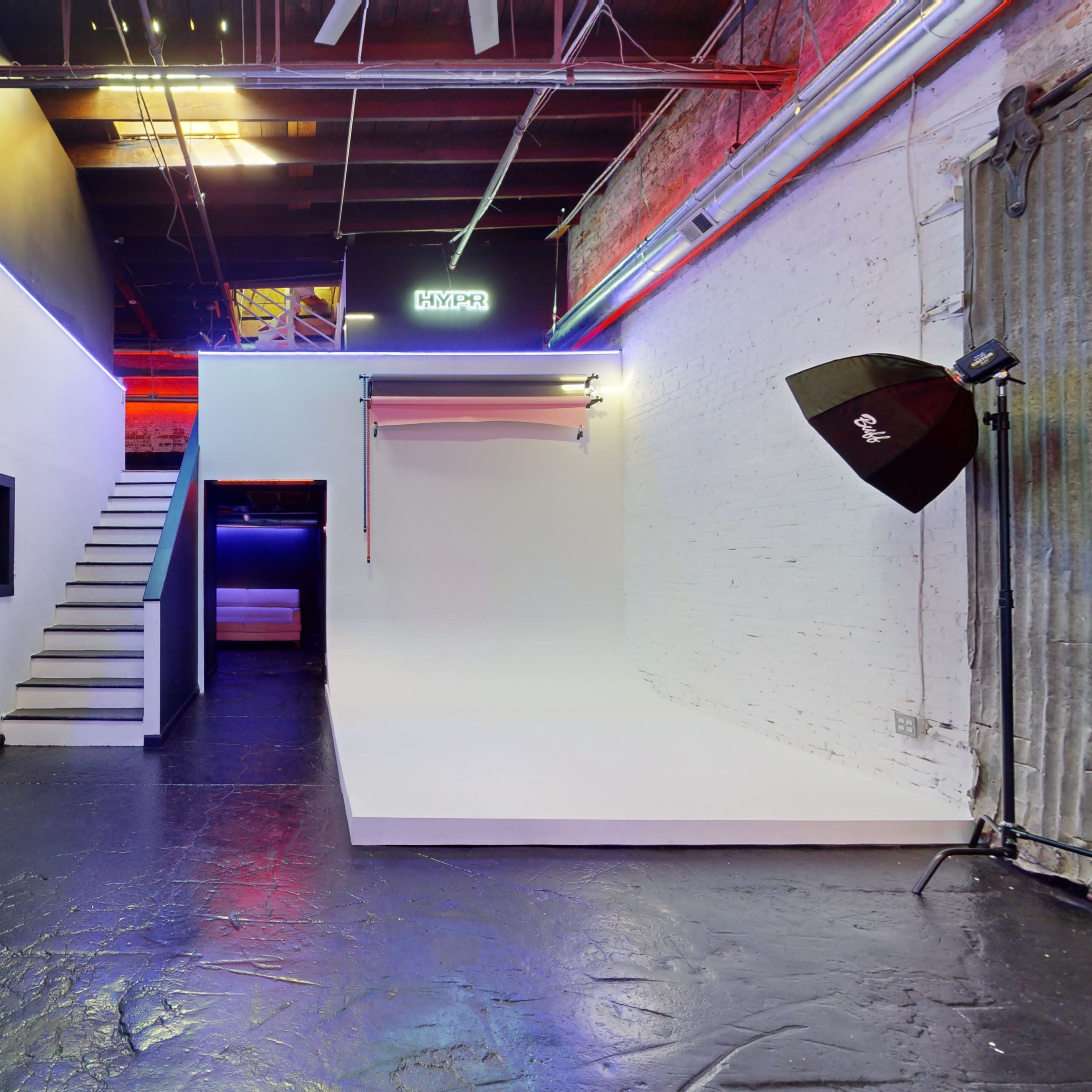 An industrial-style interior featuring a staircase leading to an upper level, a white backdrop against a wall, and studio lighting equipment.