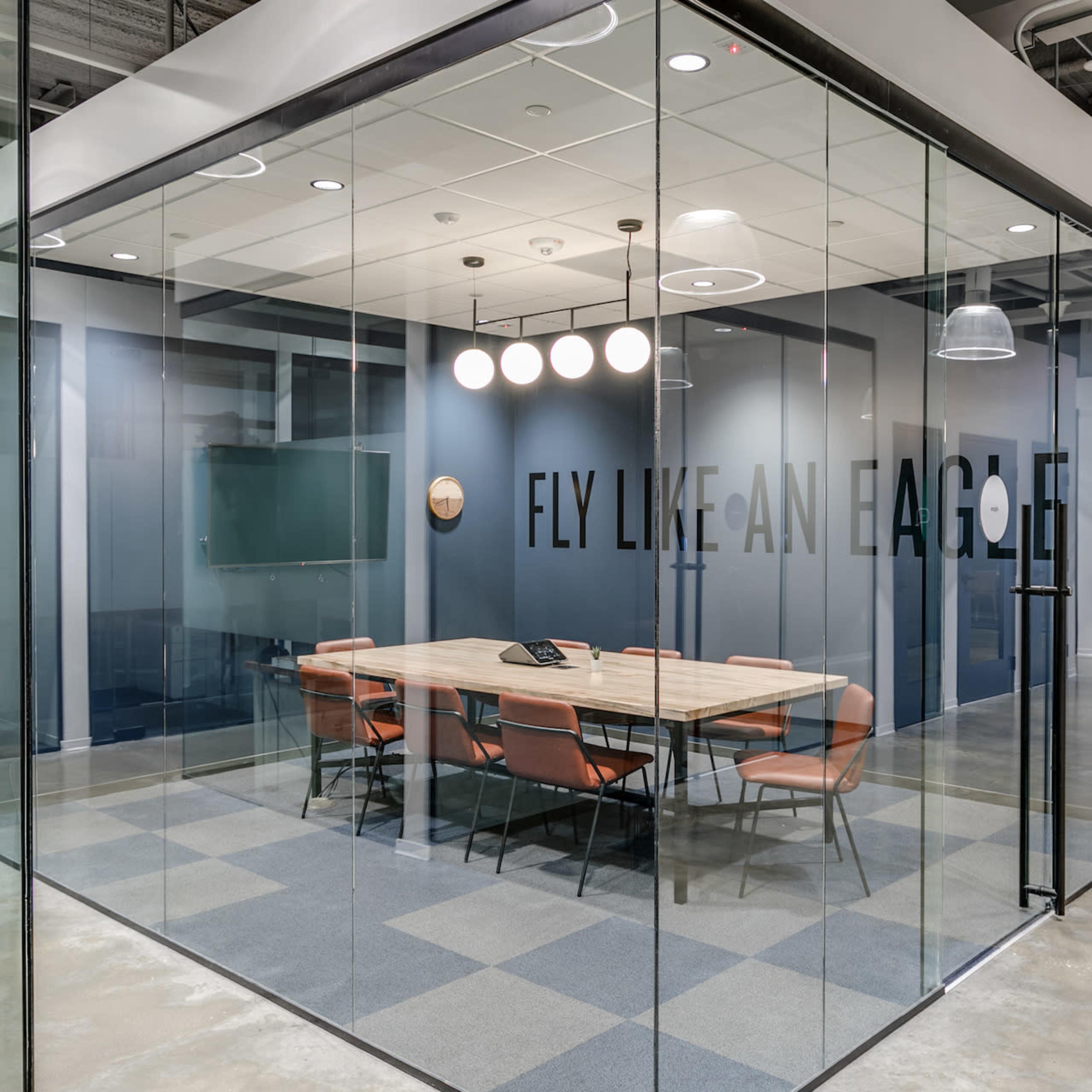 A modern conference room with a large table and orange chairs, enclosed by glass walls and featuring the phrase "FLY LIKE AN EAGLE" on one side.