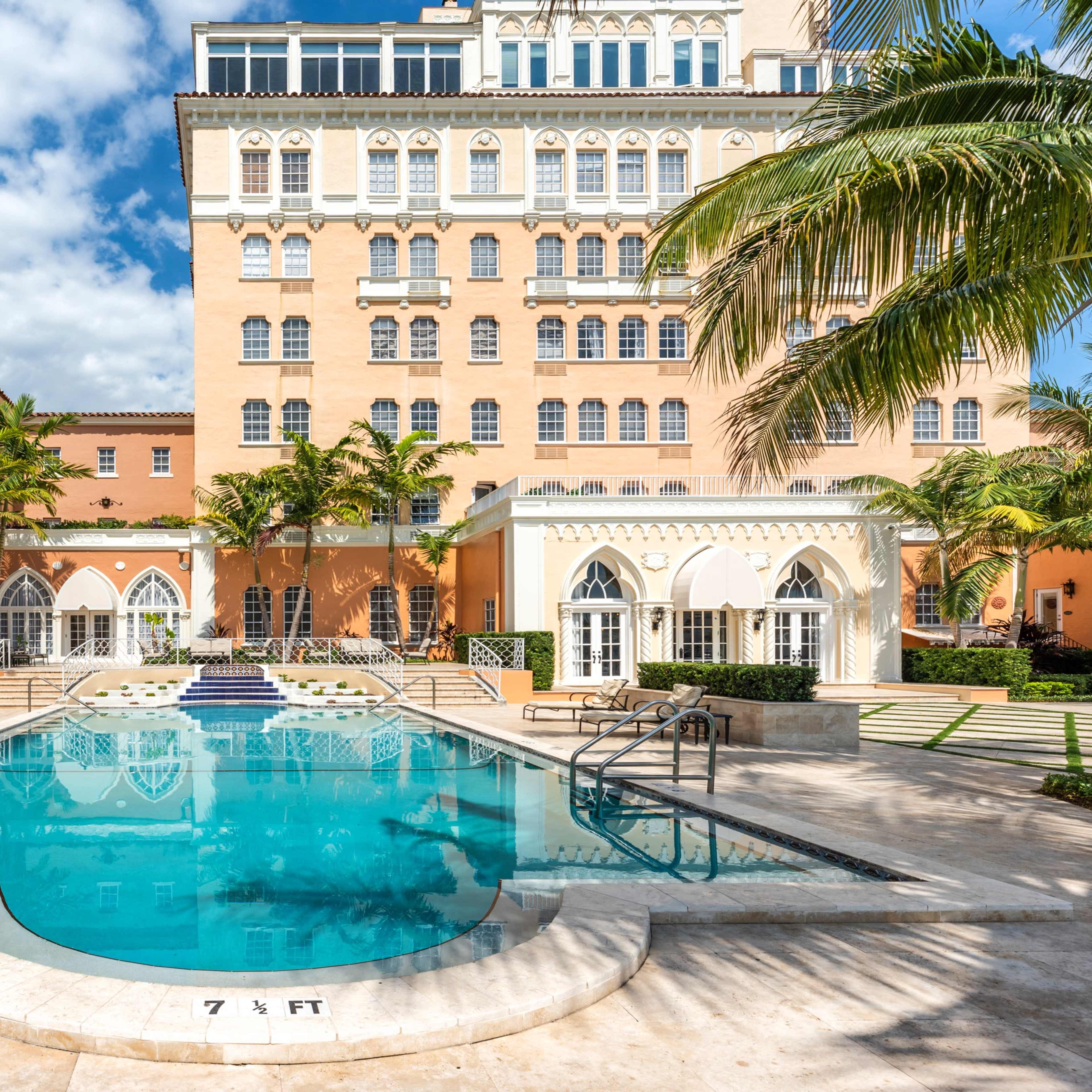 A rectangular swimming pool surrounded by palm trees and a large building with multiple windows in a courtyard setting.
