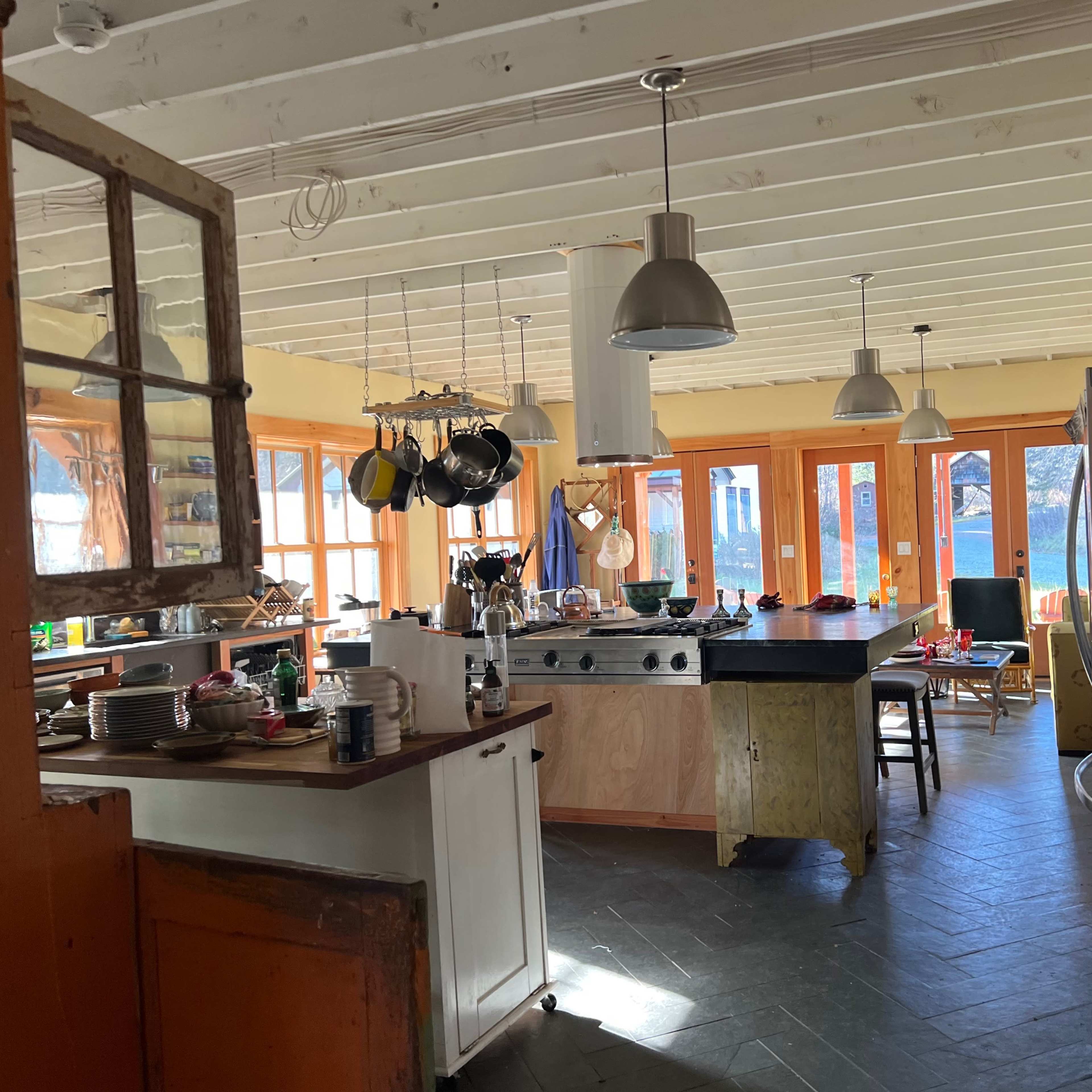 A spacious kitchen with a large island, various cooking utensils hanging from a rack, and large windows that allow natural light to enter.