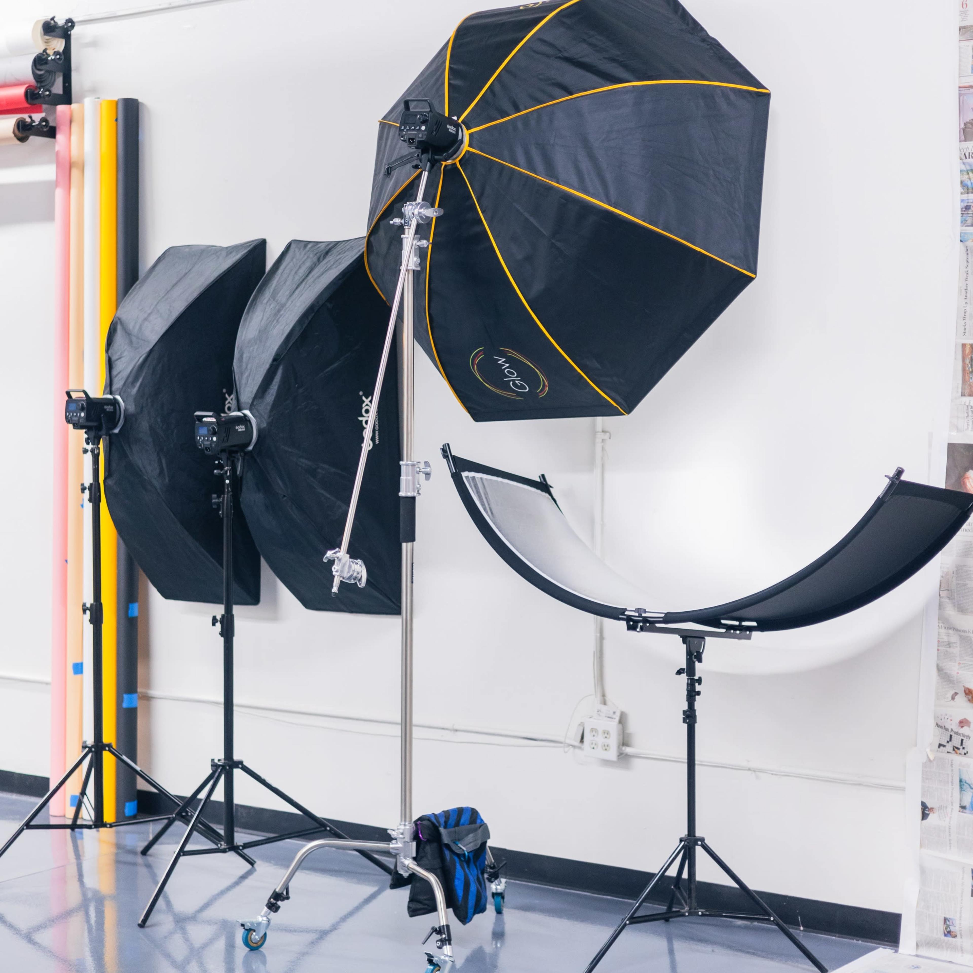 A photography studio setup with three light umbrellas and two camera tripods positioned near a backdrop support.