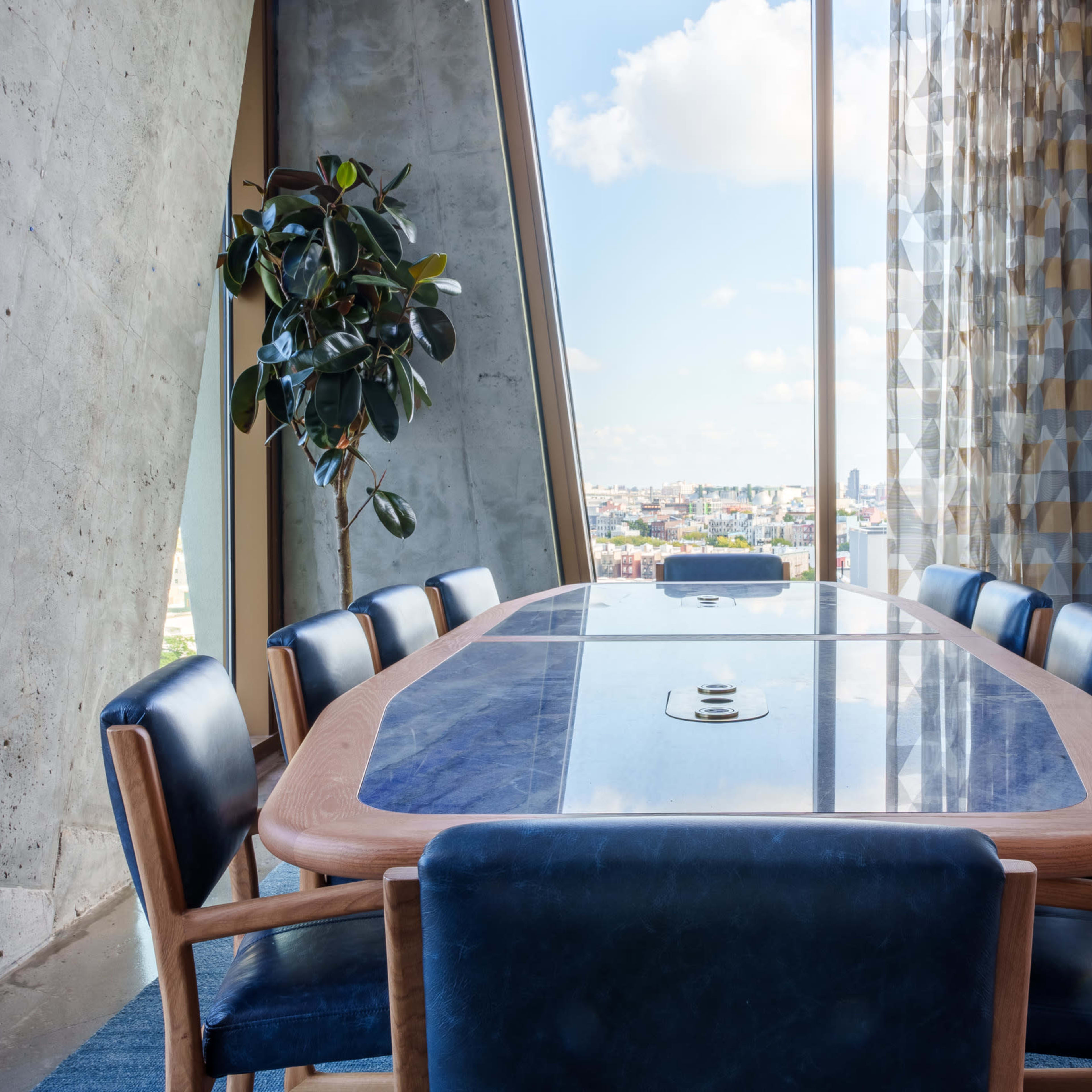 A modern conference room features a large wooden table surrounded by blue chairs, with a view of the skyline through a large window.