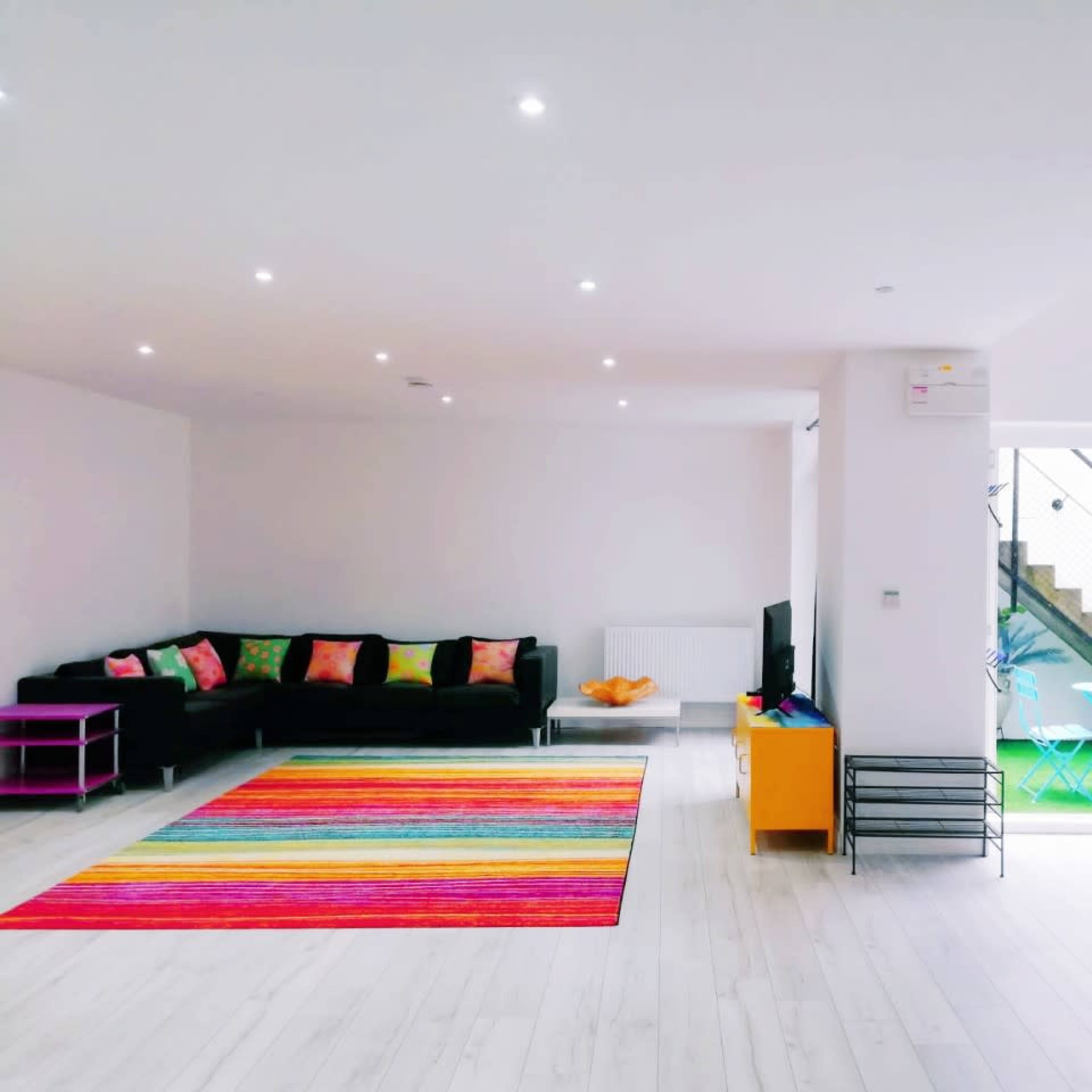 A spacious living area features a black sofa with colorful cushions, a striped rug, and a view of an outdoor patio through an open glass door.