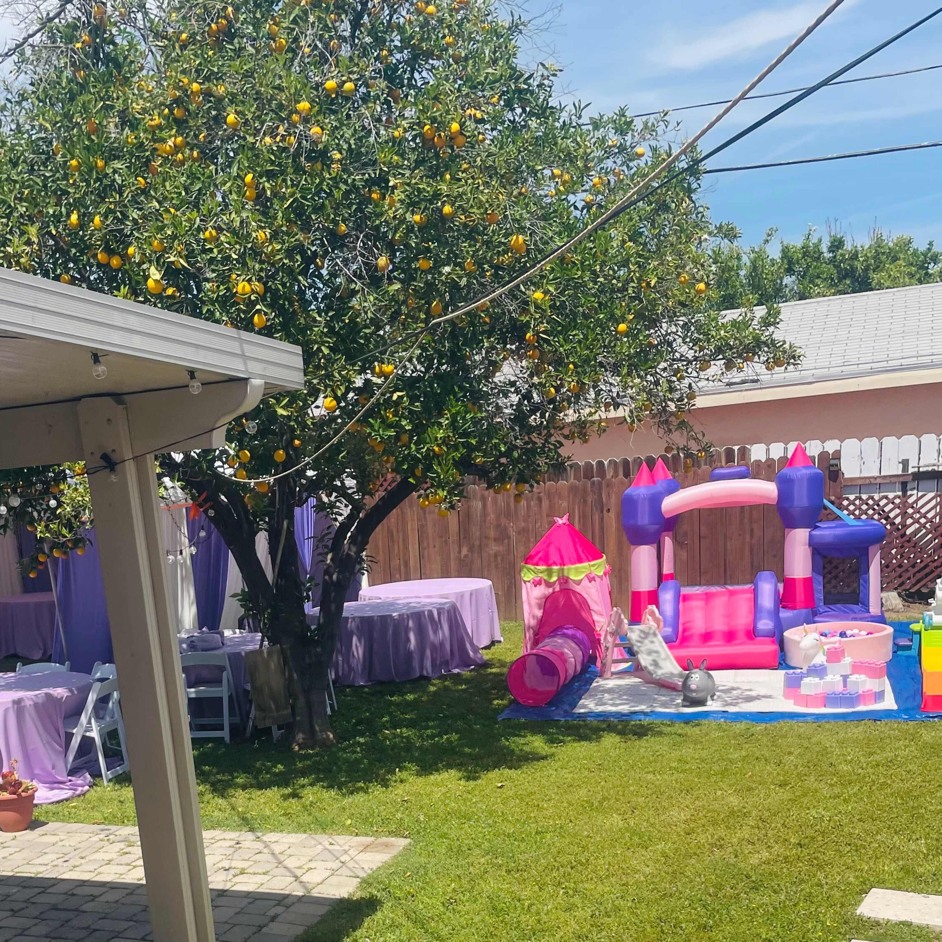 A backyard party setup features decorated tables under a tree with oranges, a pink inflatable playhouse, and a grassy area.