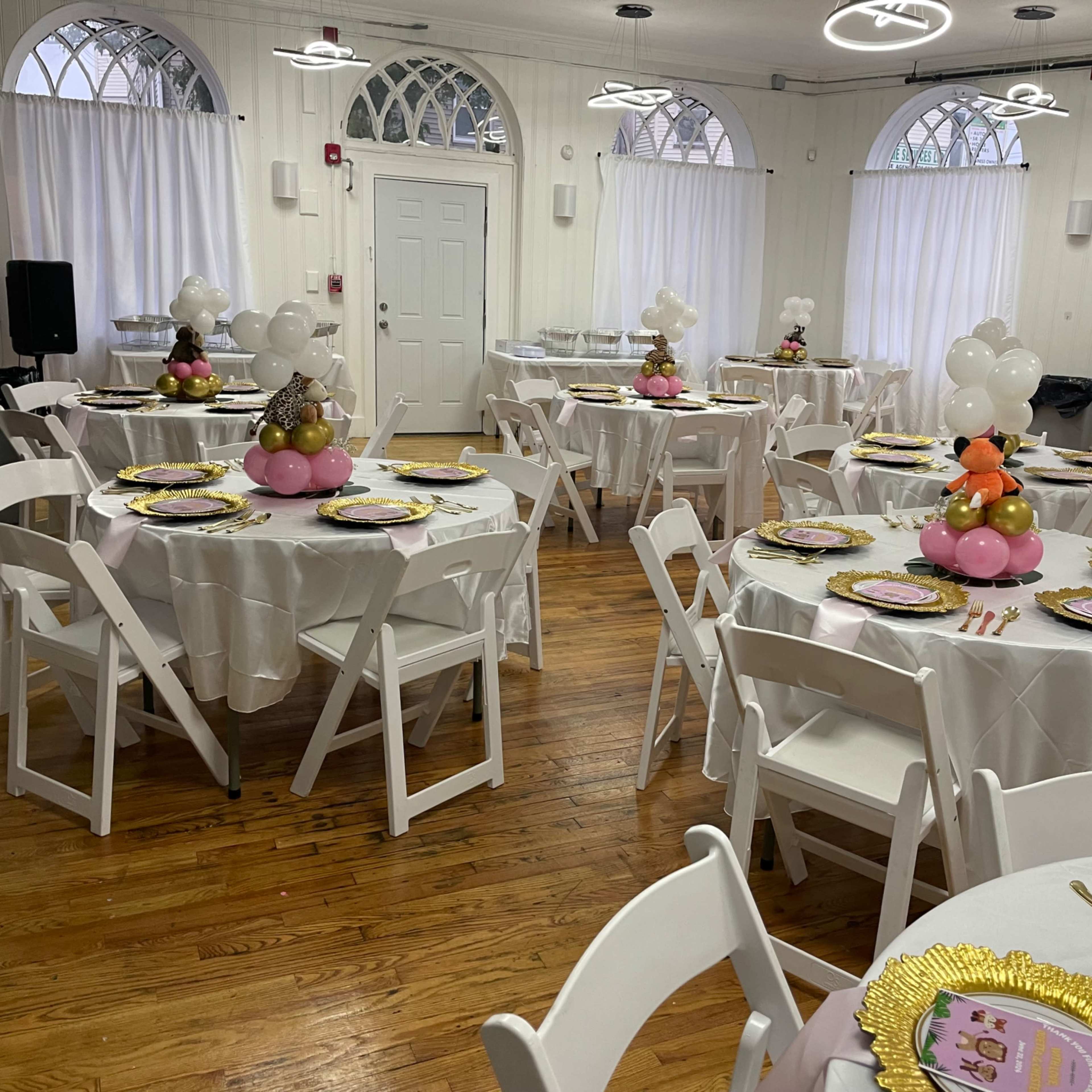 A decorated event space featuring several round tables dressed in white tablecloths, each topped with colorful centerpieces and set with gold-rimmed plates.