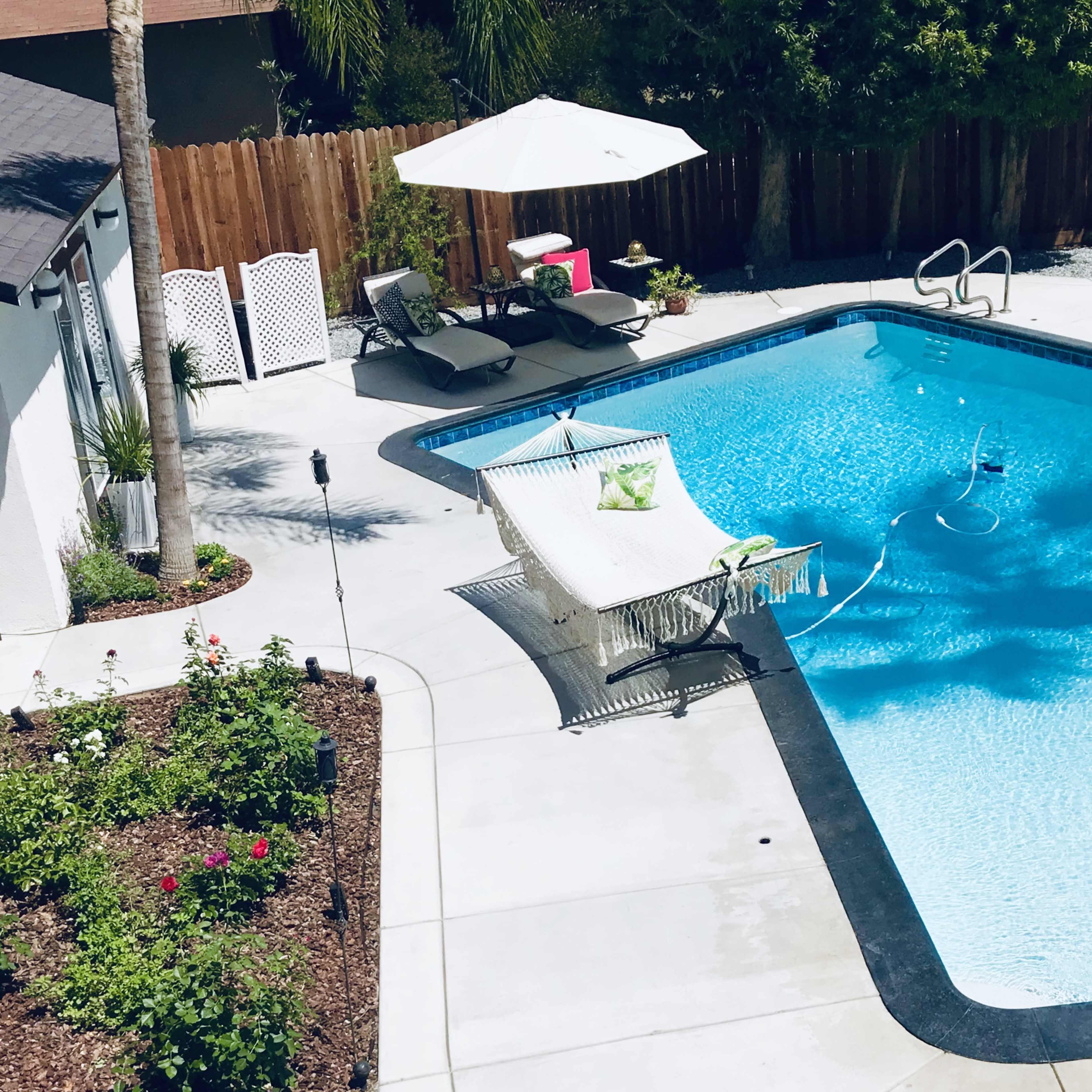 A backyard pool surrounded by lounge chairs, an umbrella, and a garden with flowers.