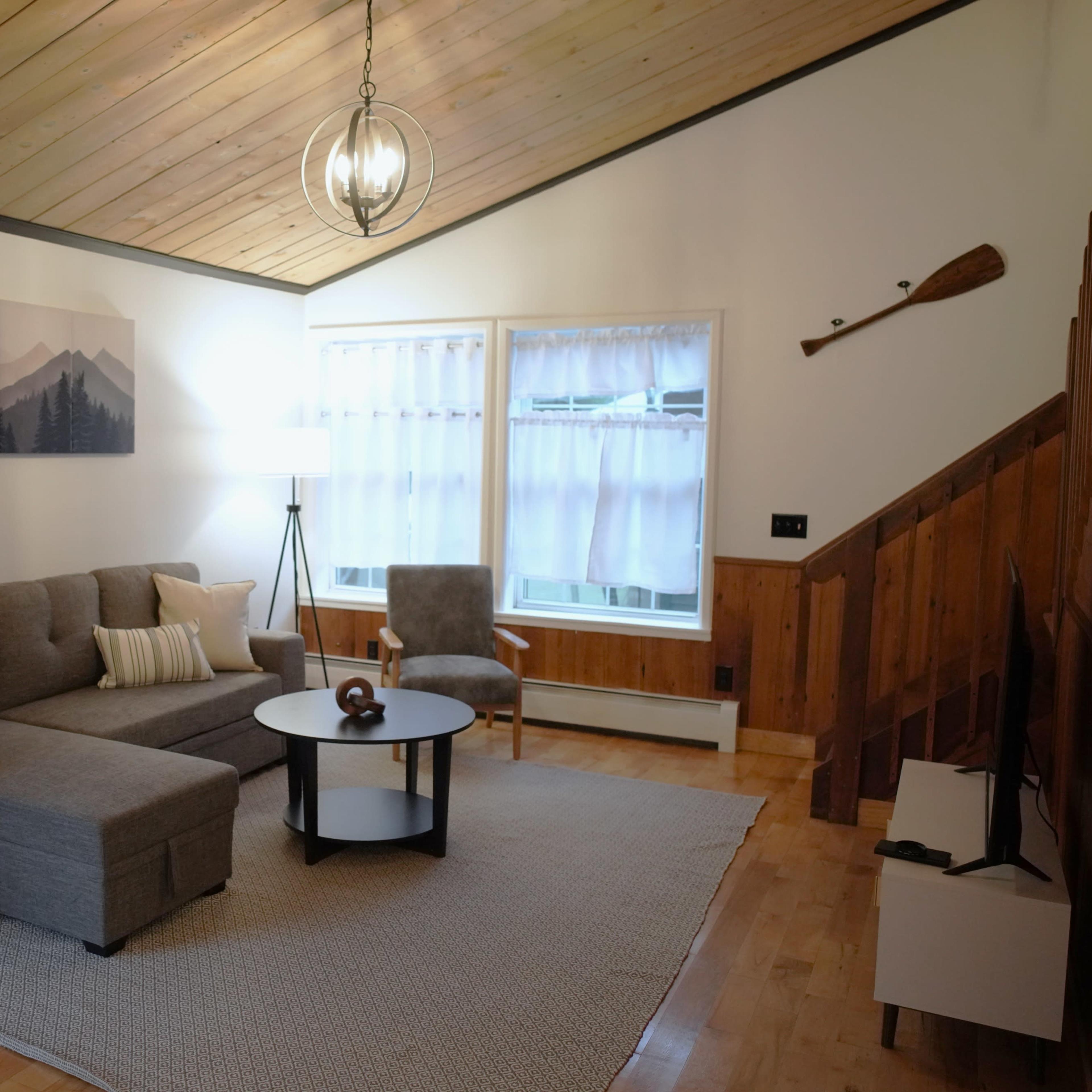 A cozy living room with a gray sectional sofa, a round coffee table, and large windows, featuring wooden accents and a high ceiling.