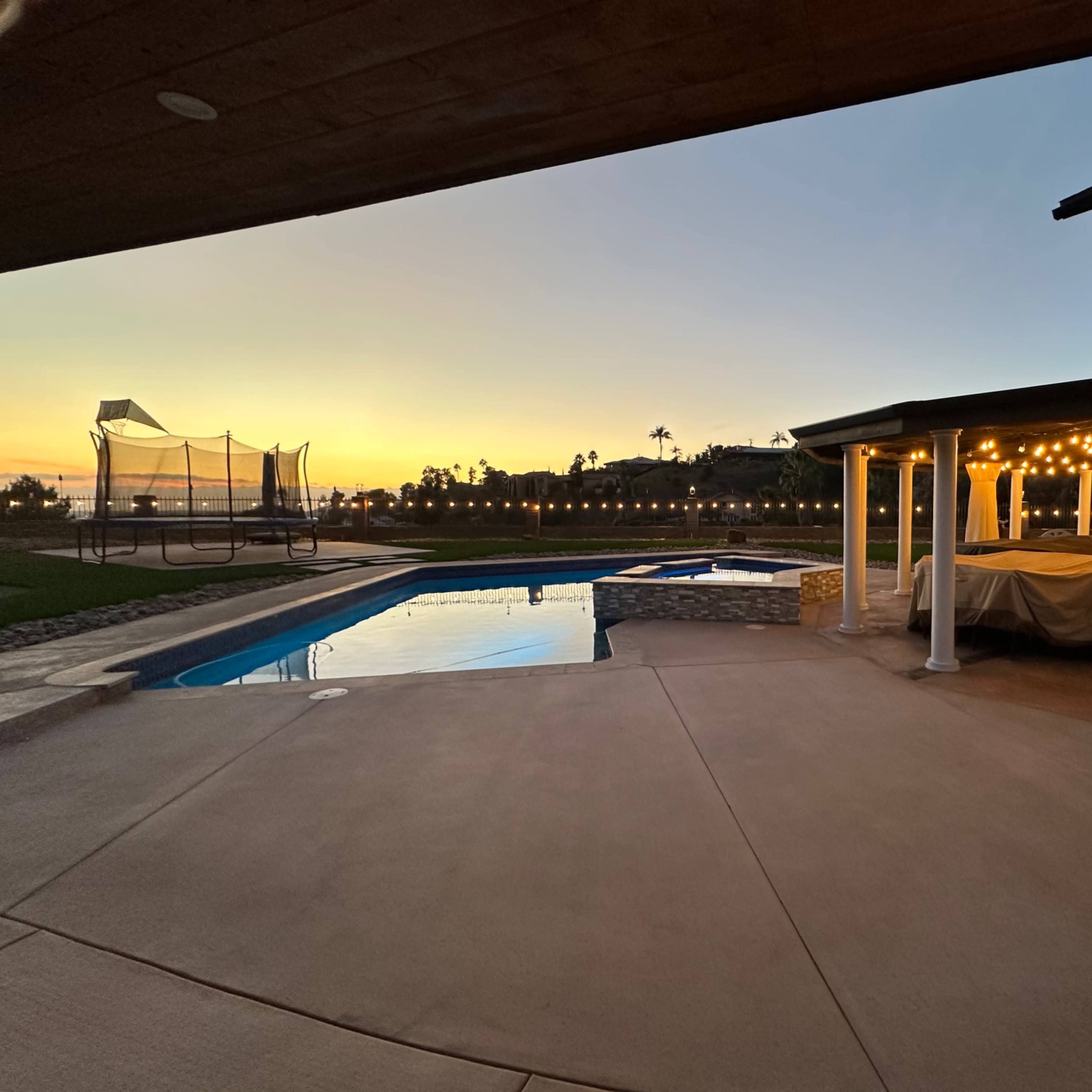 A backyard patio area with a swimming pool, a hot tub, and a covered seating space, all set against a sunset sky.