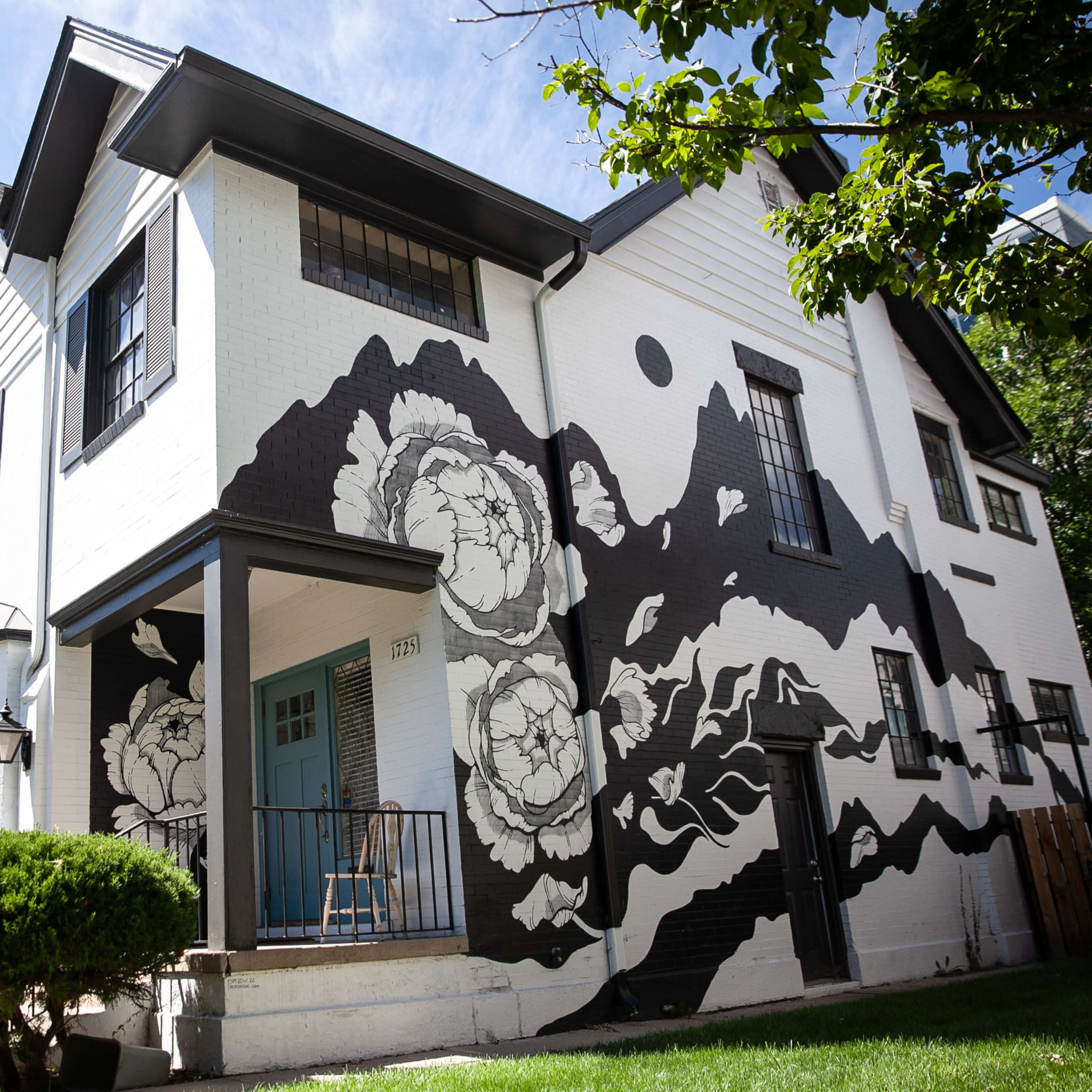 A house features a large black-and-white floral mural on one side, with a front porch and well-maintained landscaping.