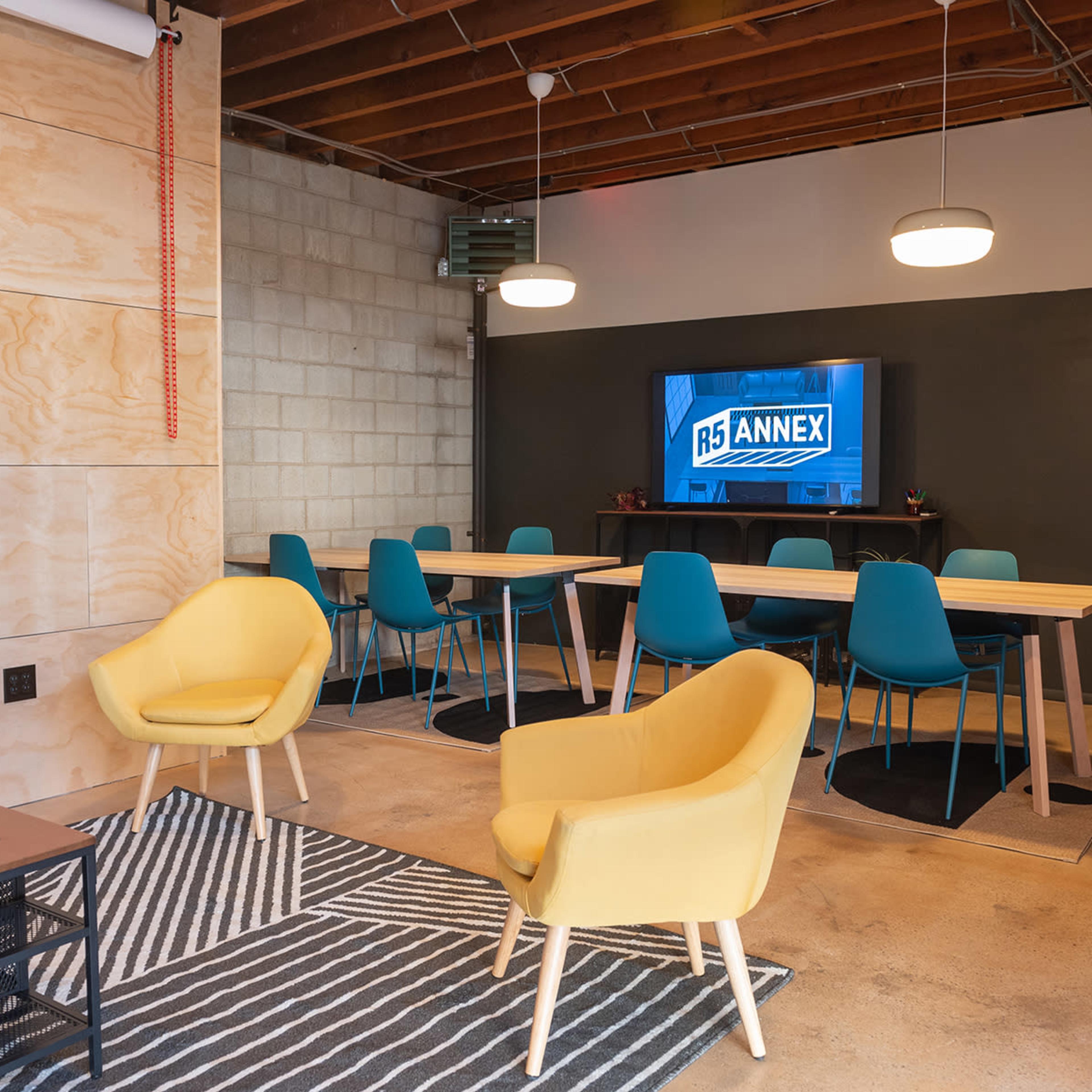 A modern meeting room features a long table surrounded by teal chairs, two yellow armchairs, and a television displaying the "R5 Annex" logo.