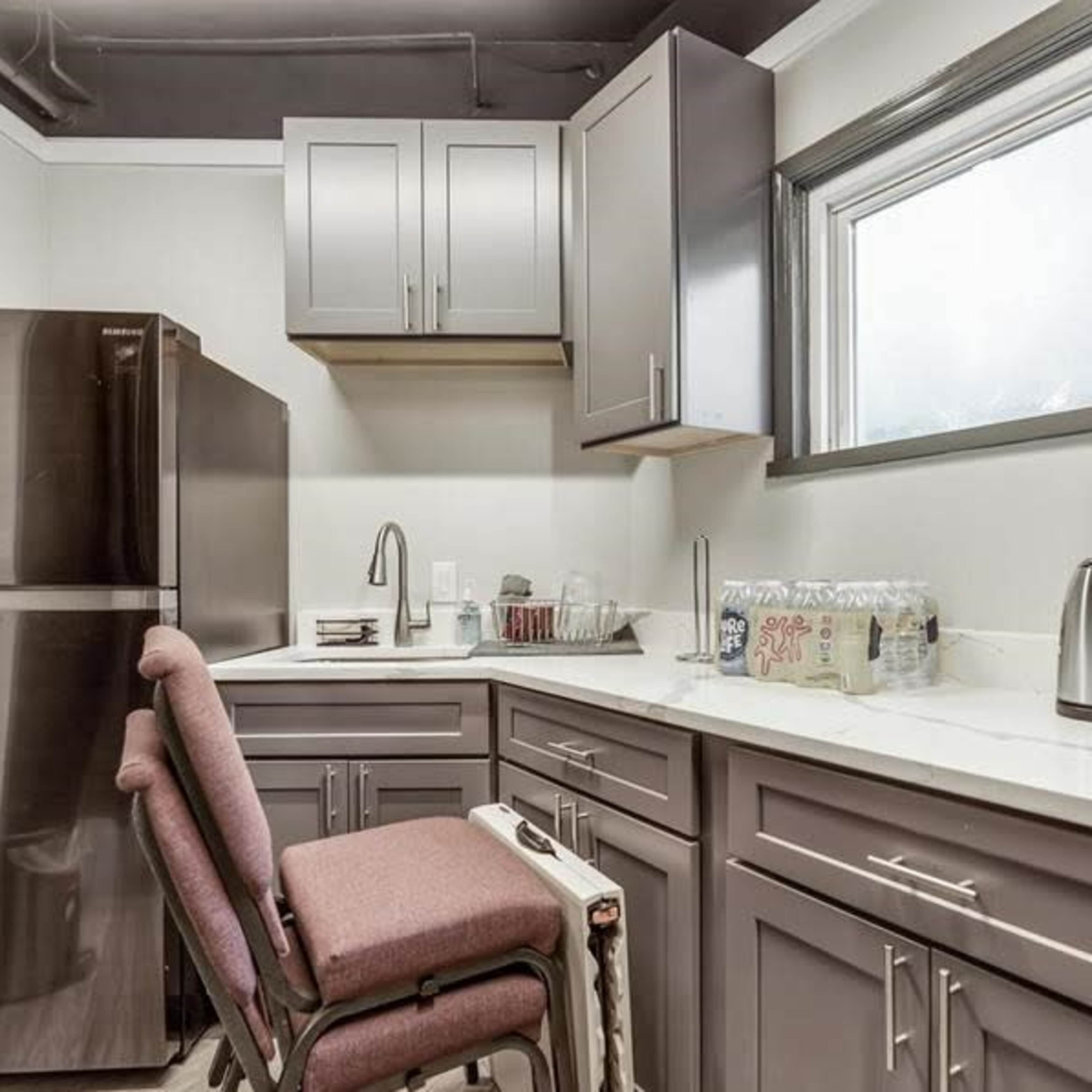 A small kitchen area with gray cabinets, a refrigerator, a sink, and a countertop, accompanied by a chair and kitchen appliances.