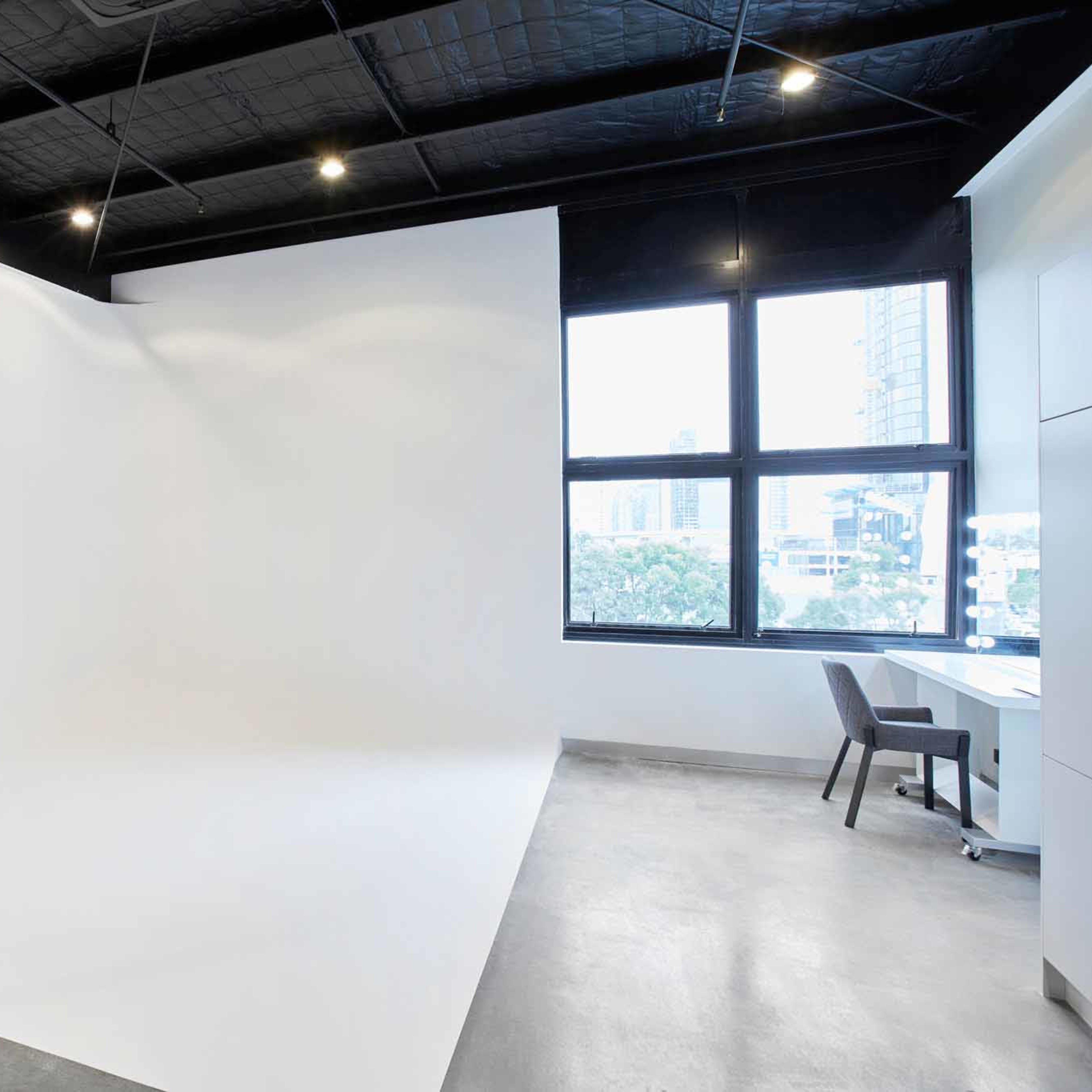 A spacious photography studio with a white backdrop, large windows, and a simple desk with a chair.