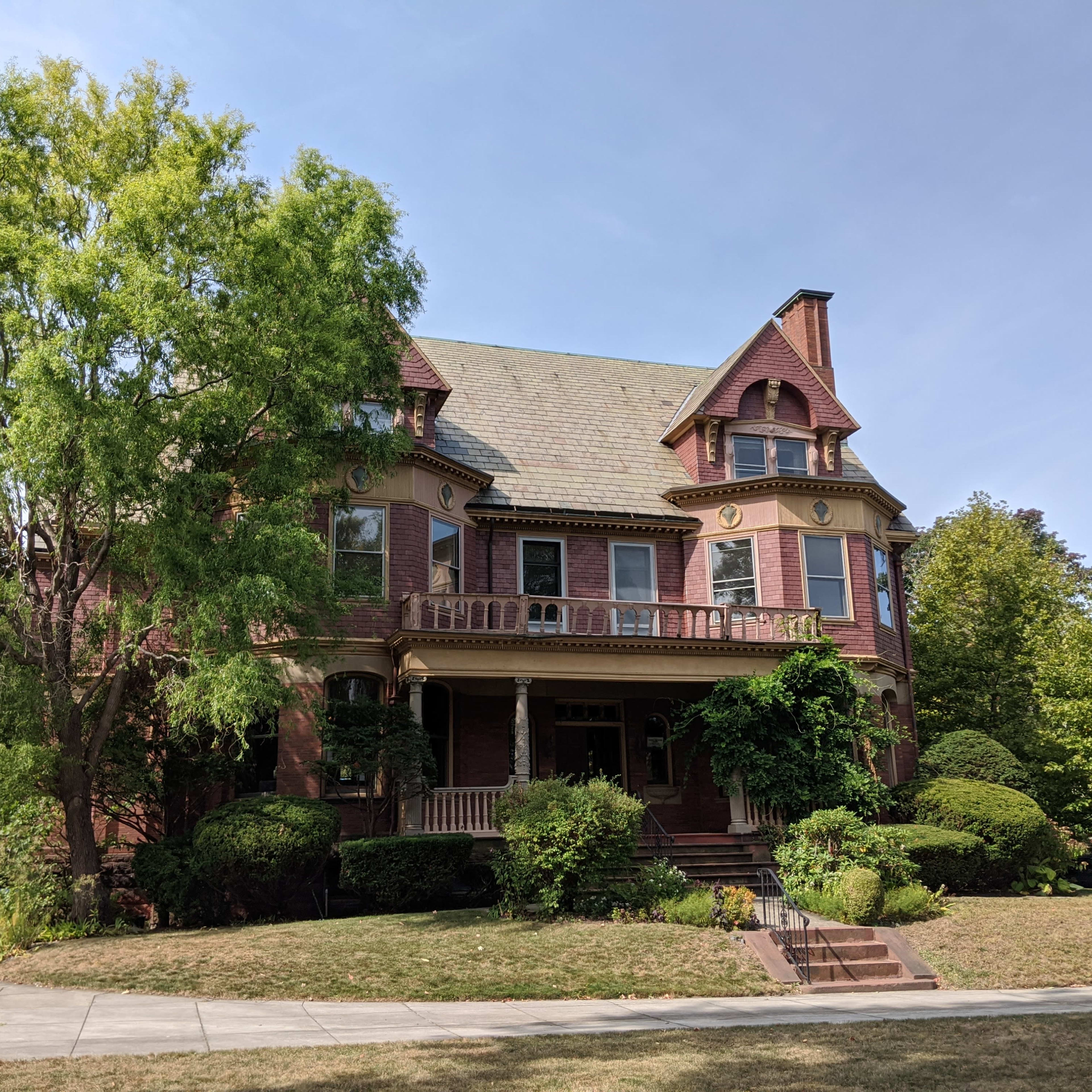 A large, red brick Victorian house stands surrounded by lush greenery and well-maintained shrubs on a clear day.