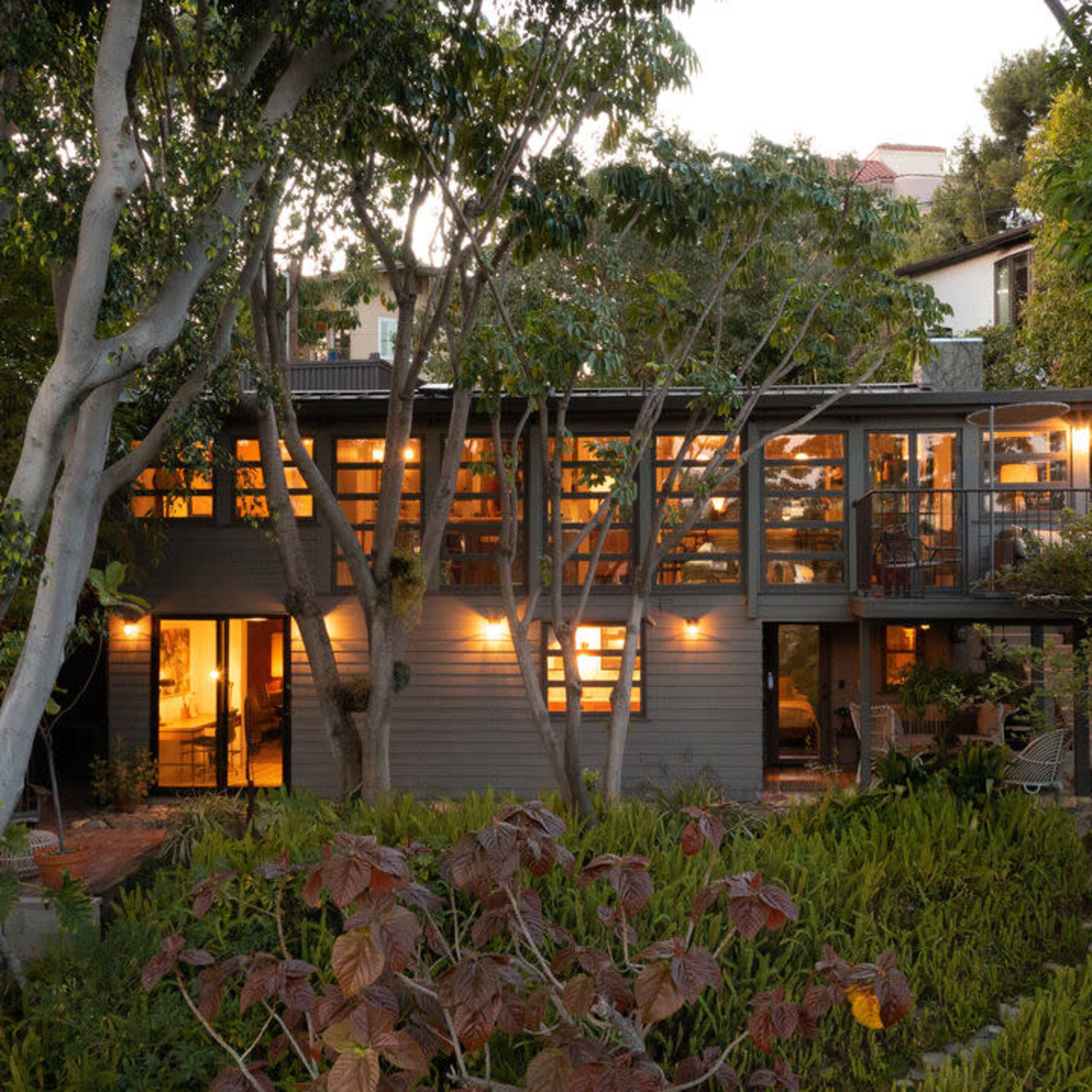 A two-story, modern residential house features large windows and a deck, surrounded by landscaped greenery and trees.