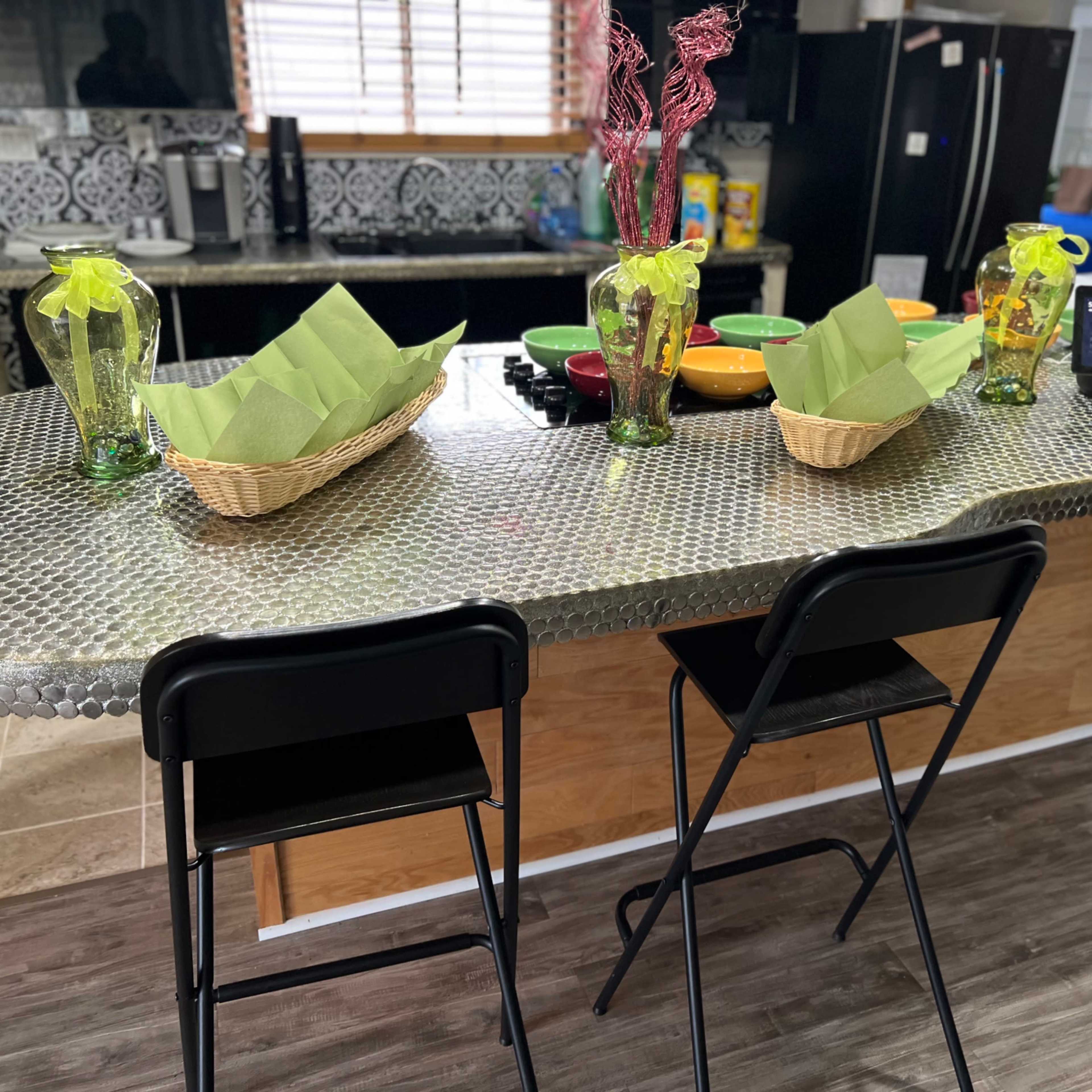 A kitchen bar area with a metallic countertop, two black stools, and decorative items including vases and baskets with green napkins.