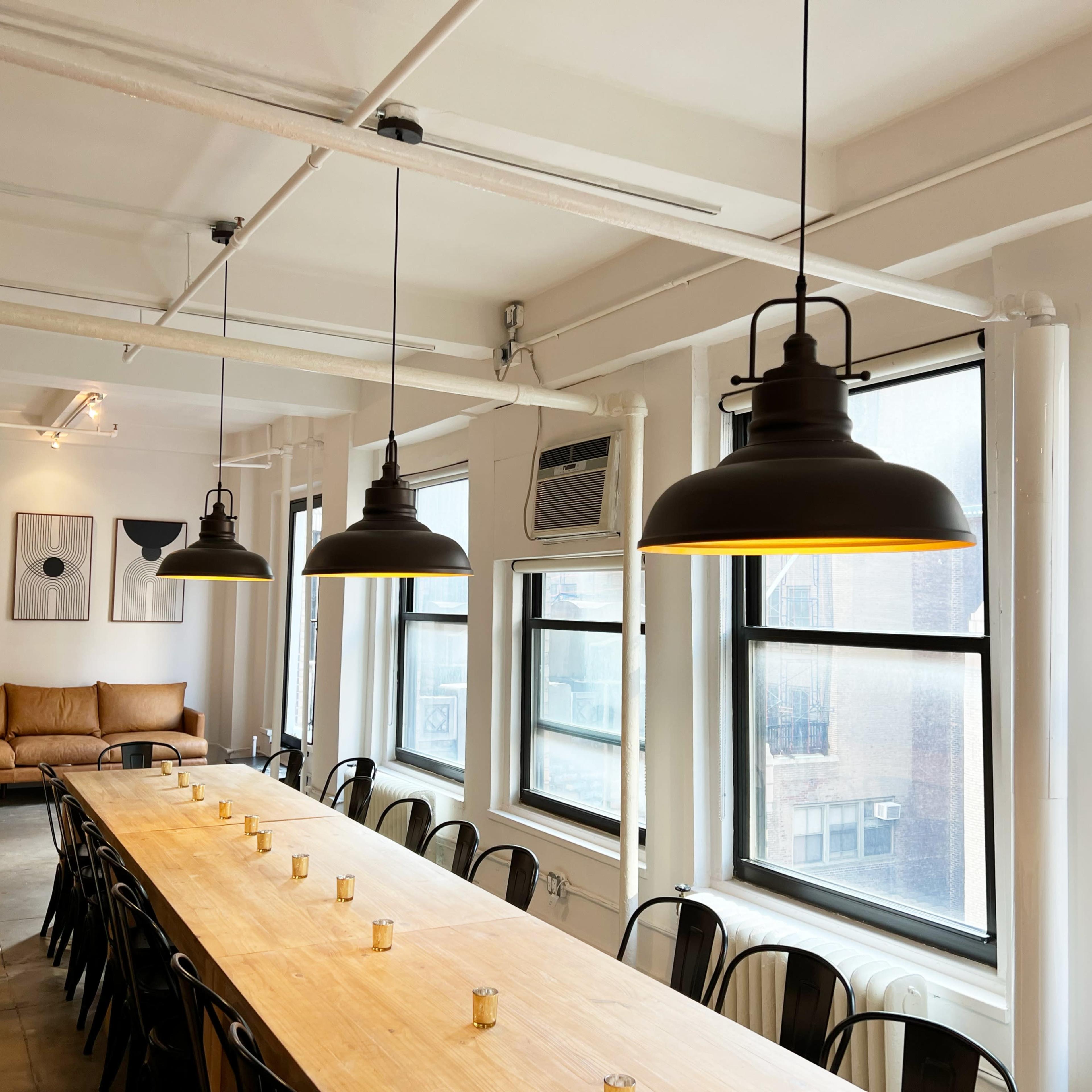 A long wooden table is set under industrial-style pendant lights in a well-lit room with windows, a couch, and a Christmas tree in the corner.