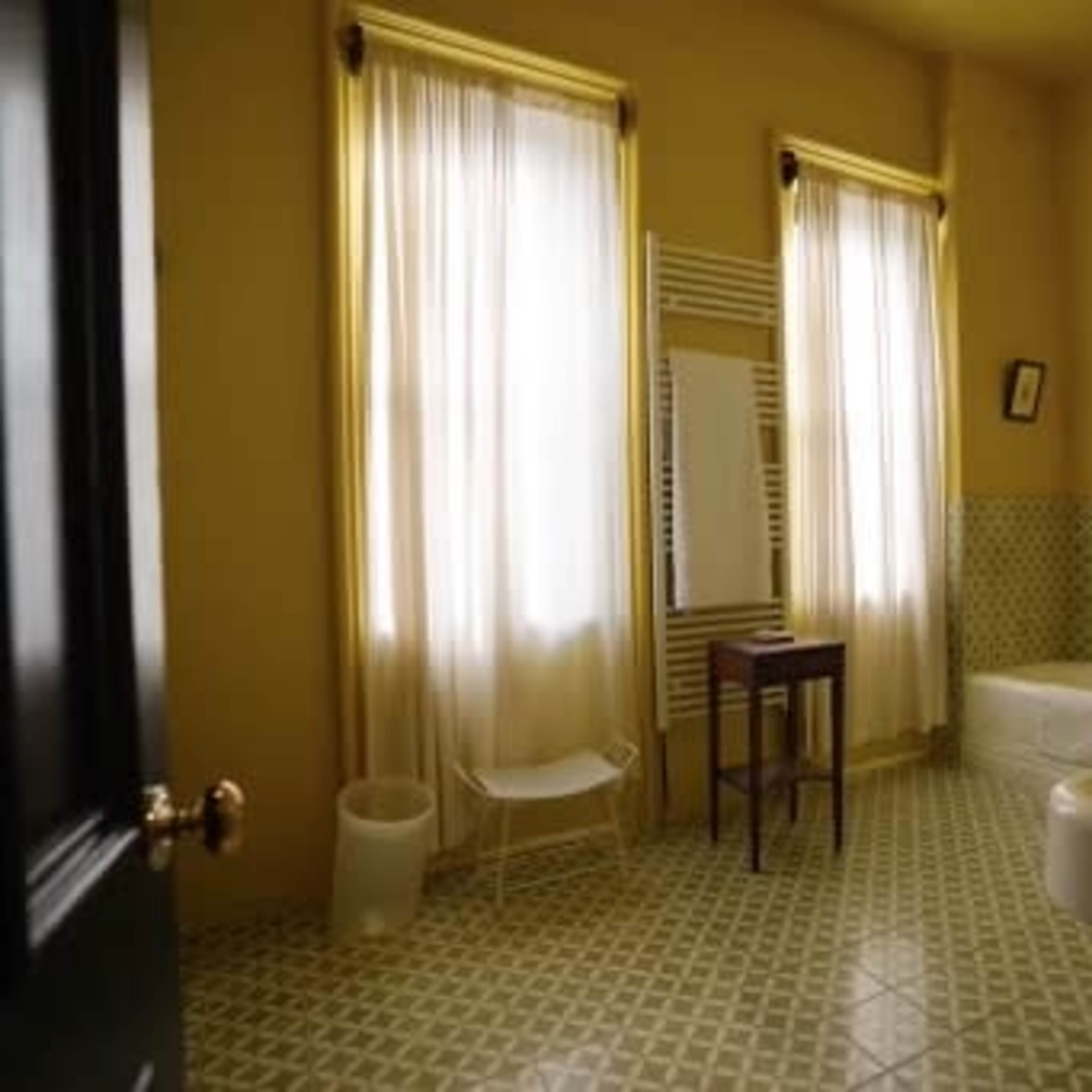 The scene depicts a yellow bathroom featuring a bathtub, a small wooden table, a mirror, and large windows with sheer curtains.